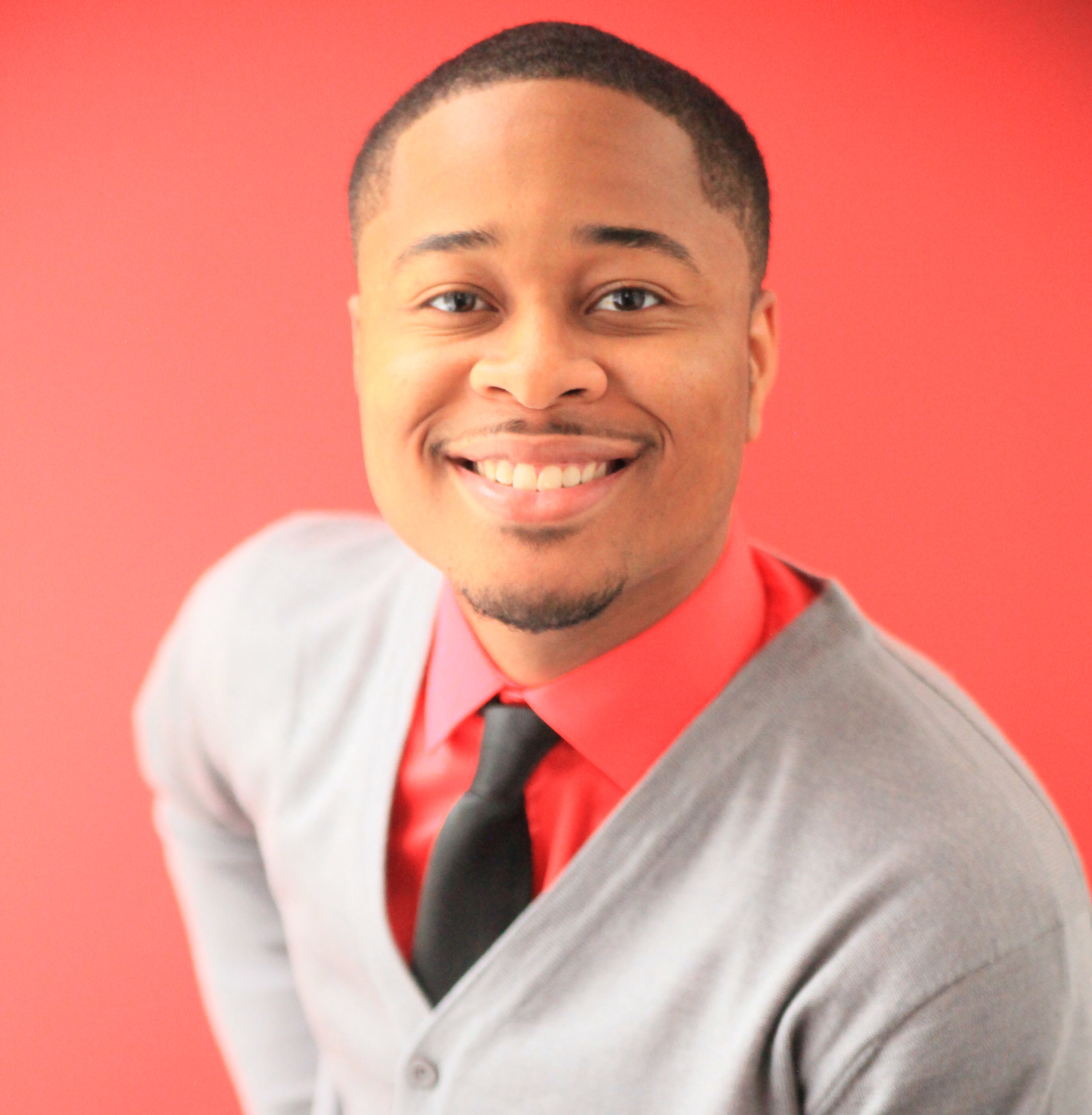 Portrait of a smiling man wearing a gray blazer, red shirt, and black tie against a red background.