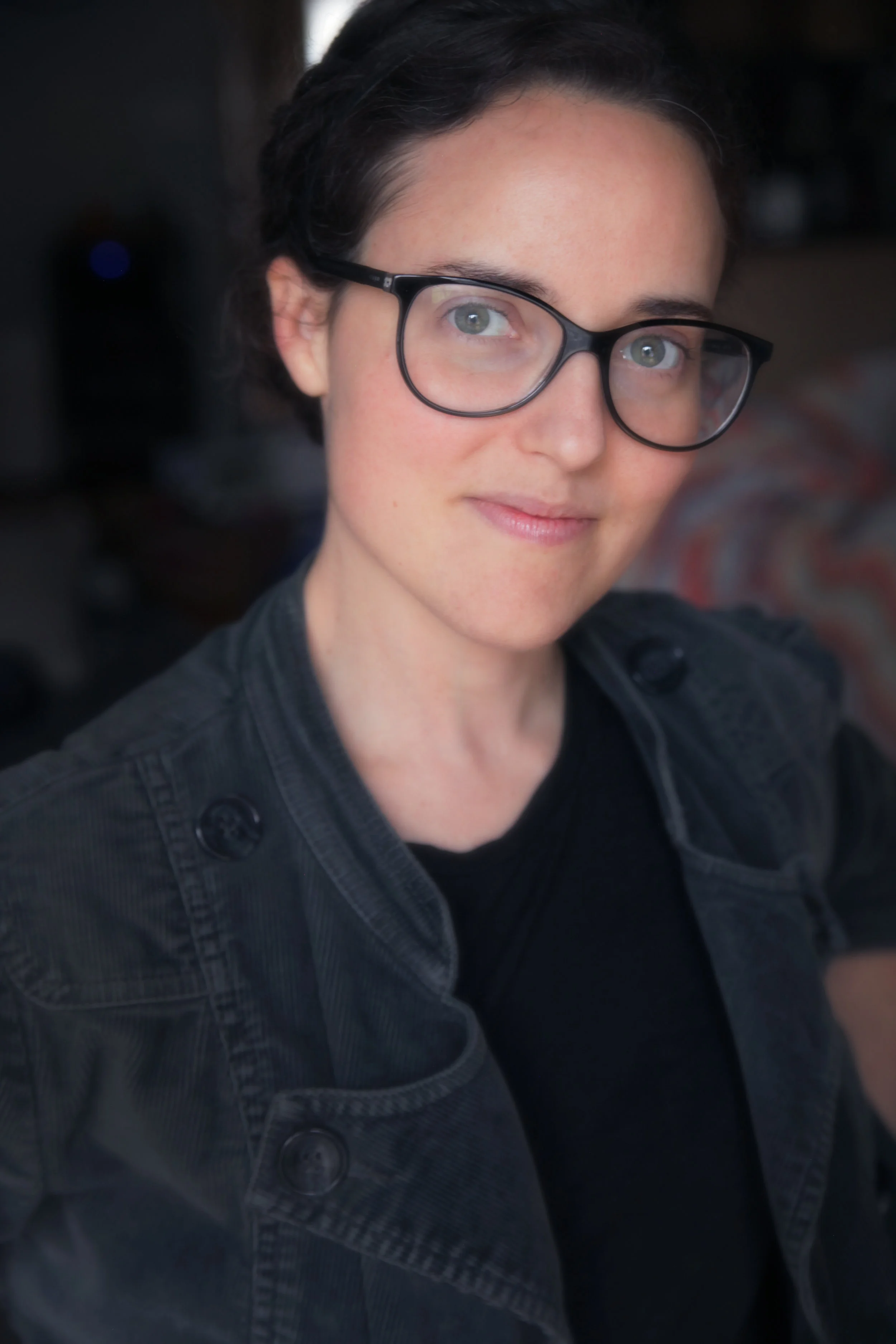 Close-up of a woman with short dark hair wearing glasses and a black jacket, smiling slightly indoors.