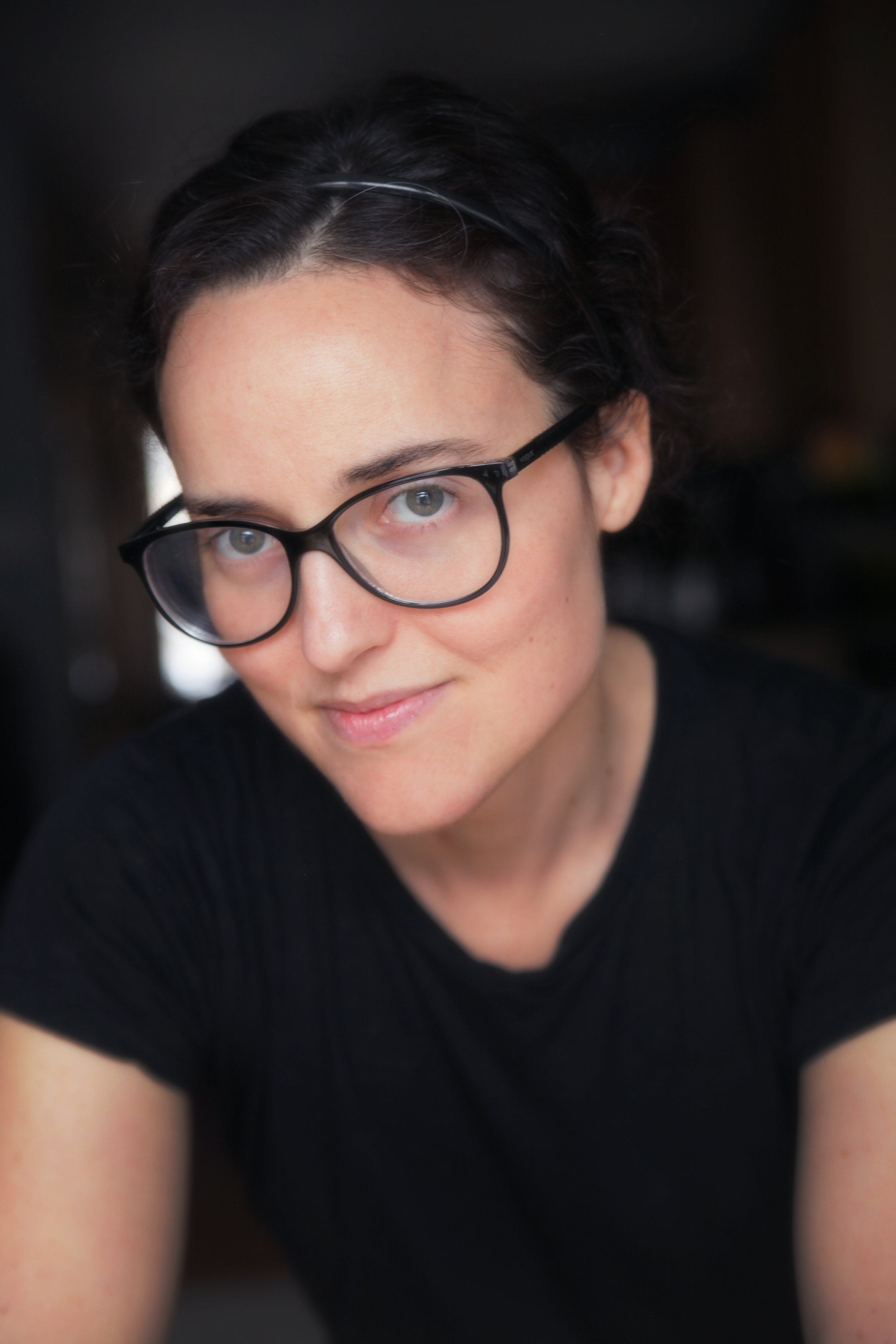 Close-up portrait of a woman with short dark hair, glasses, and a black shirt, smiling softly at the camera.