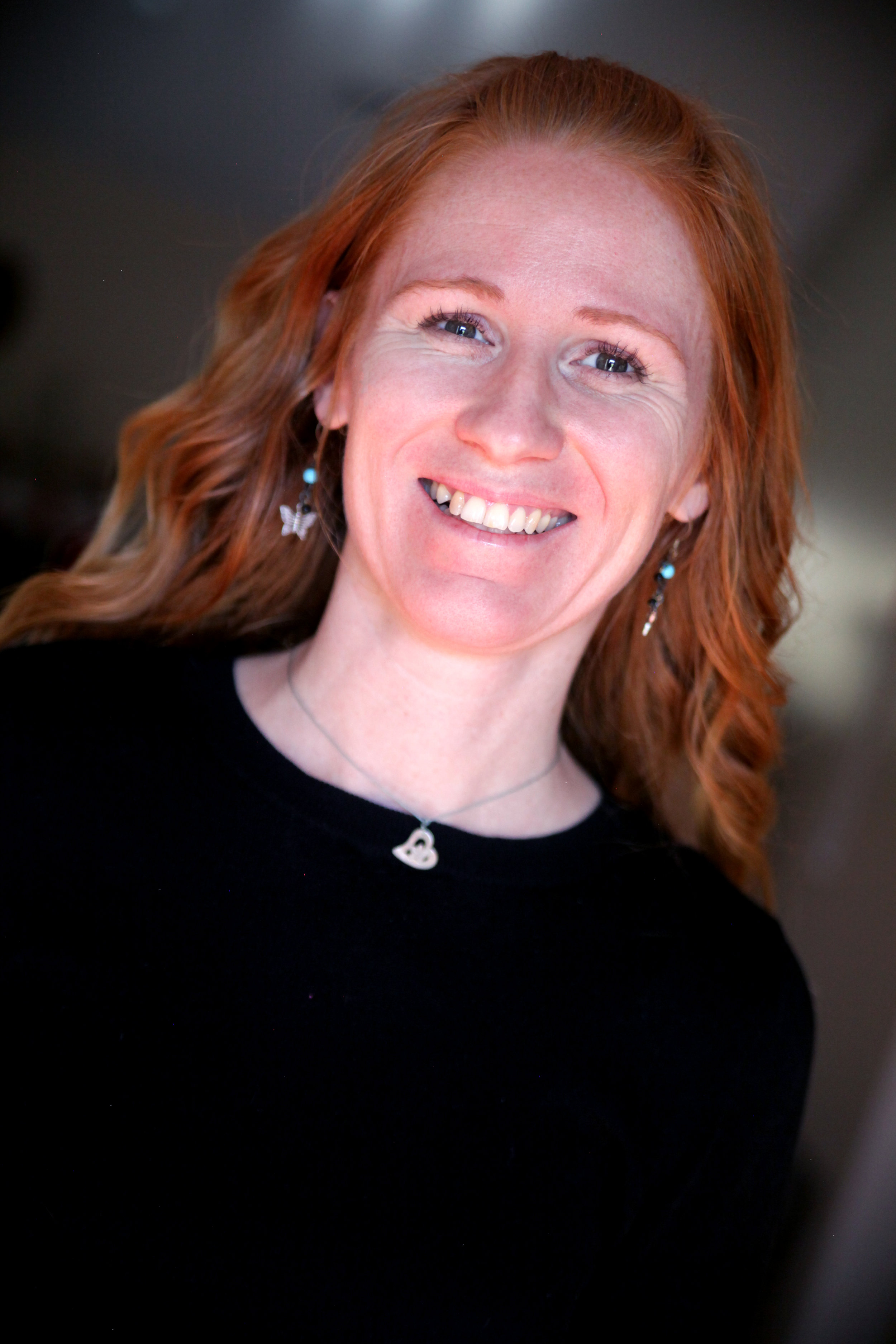 A woman with red hair smiling, wearing earrings and a necklace with a heart pendant, dressed in a black top.