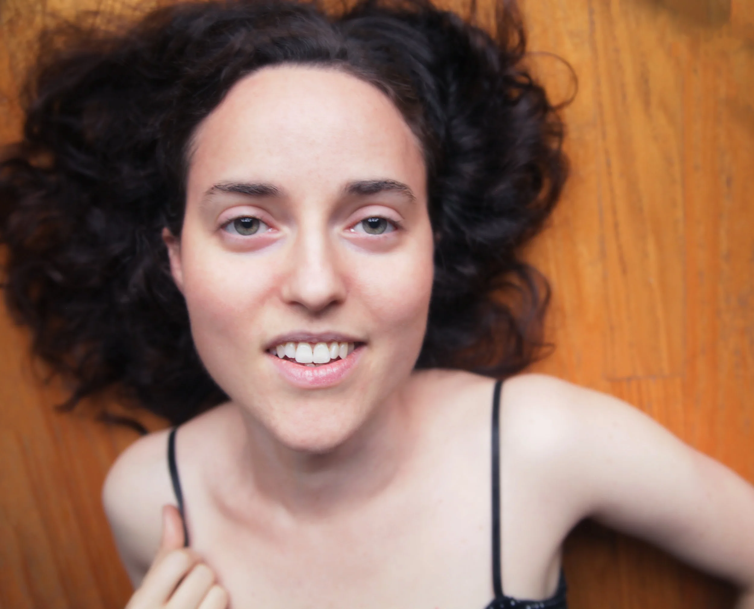 A woman with curly dark hair lying down on a wooden floor, smiling at the camera, wearing a black spaghetti strap top.