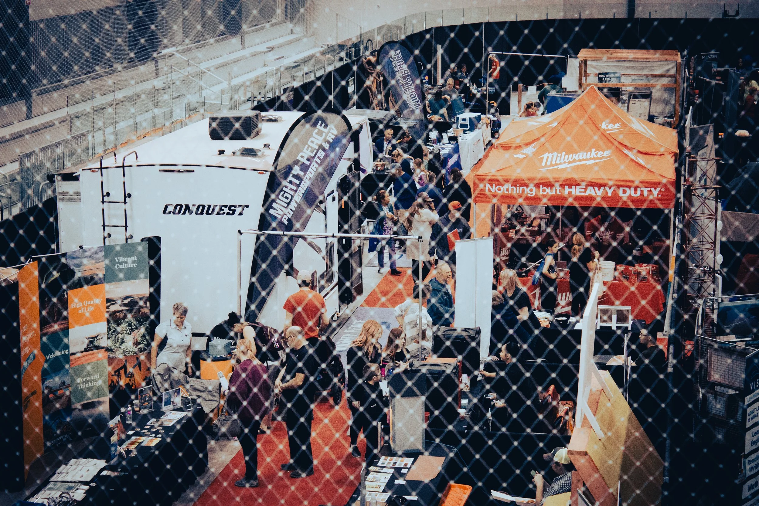 View through arena netting of people milling an aisle at a trade show, there is an RV to the left and a tent booth to the right.