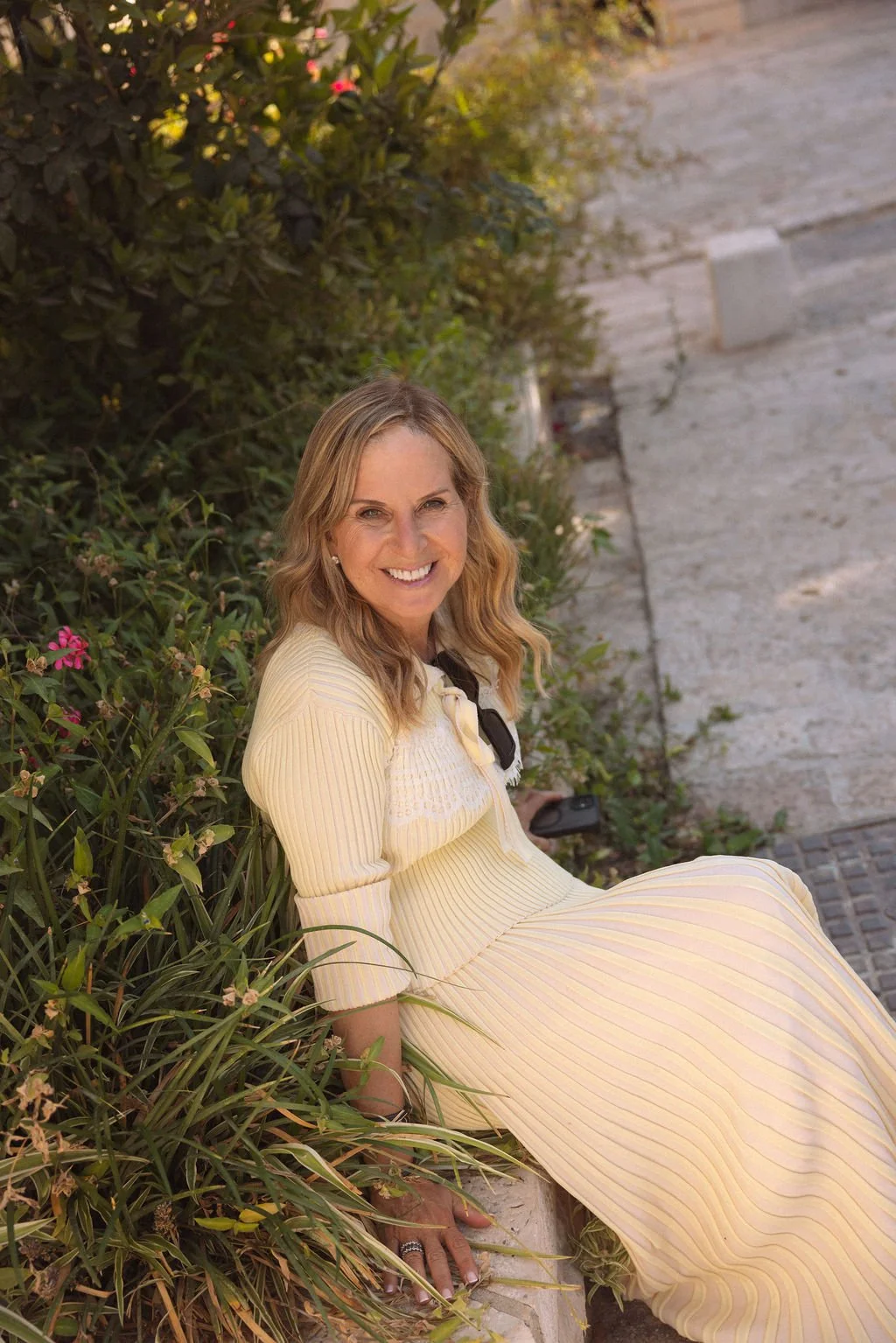 A woman with blonde hair wearing a yellow and white striped dress, sitting on the sidewalk next to green bushes with pink flowers, smiling at the camera.