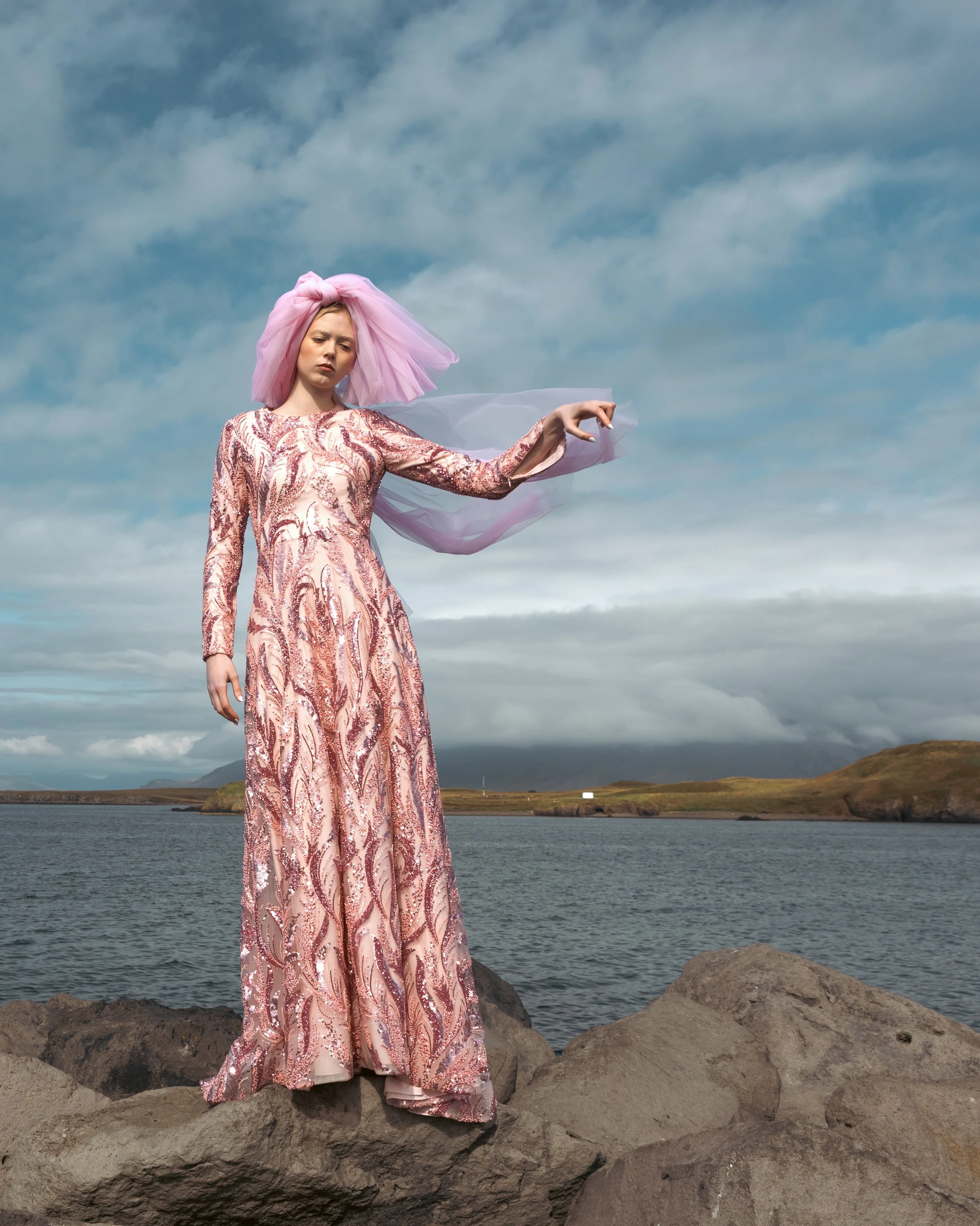 A woman standing on rocks near a body of water, wearing a long, elegant pink and gold dress with a matching pink tulle headwrap, holding her arm out against a cloudy sky and scenic landscape.