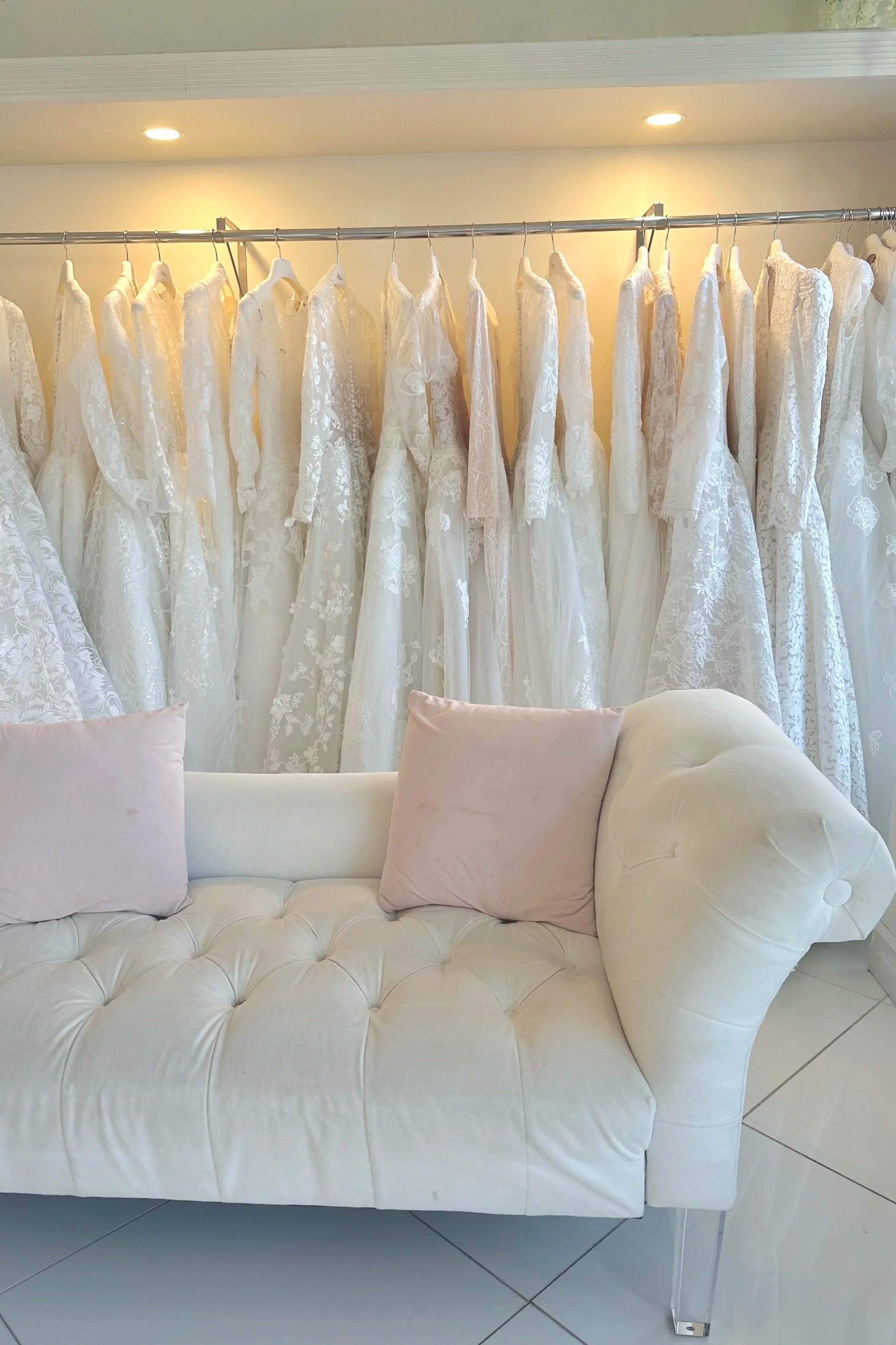 White bridal dresses hanging on a rack behind a white sofa with two pink pillows in a bridal boutique or fitting room.