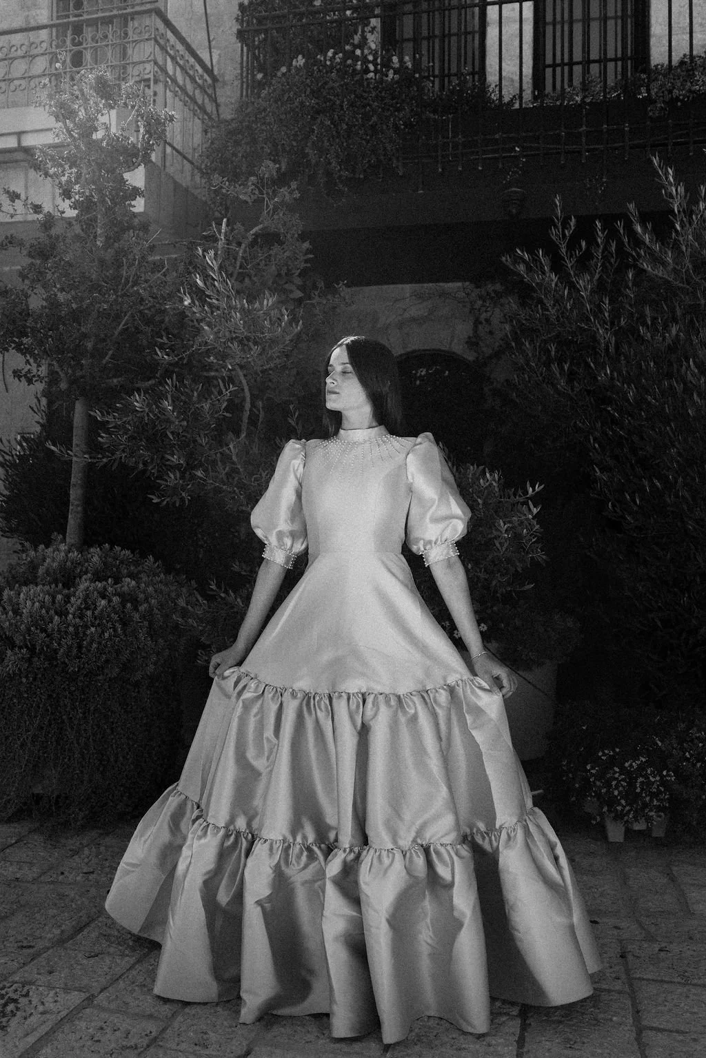 A woman with dark hair in an elegant, vintage-style satin gown with puffed sleeves and a ruffled hem, standing outdoors at night among lush bushes and trees, with a building and balcony in the background.