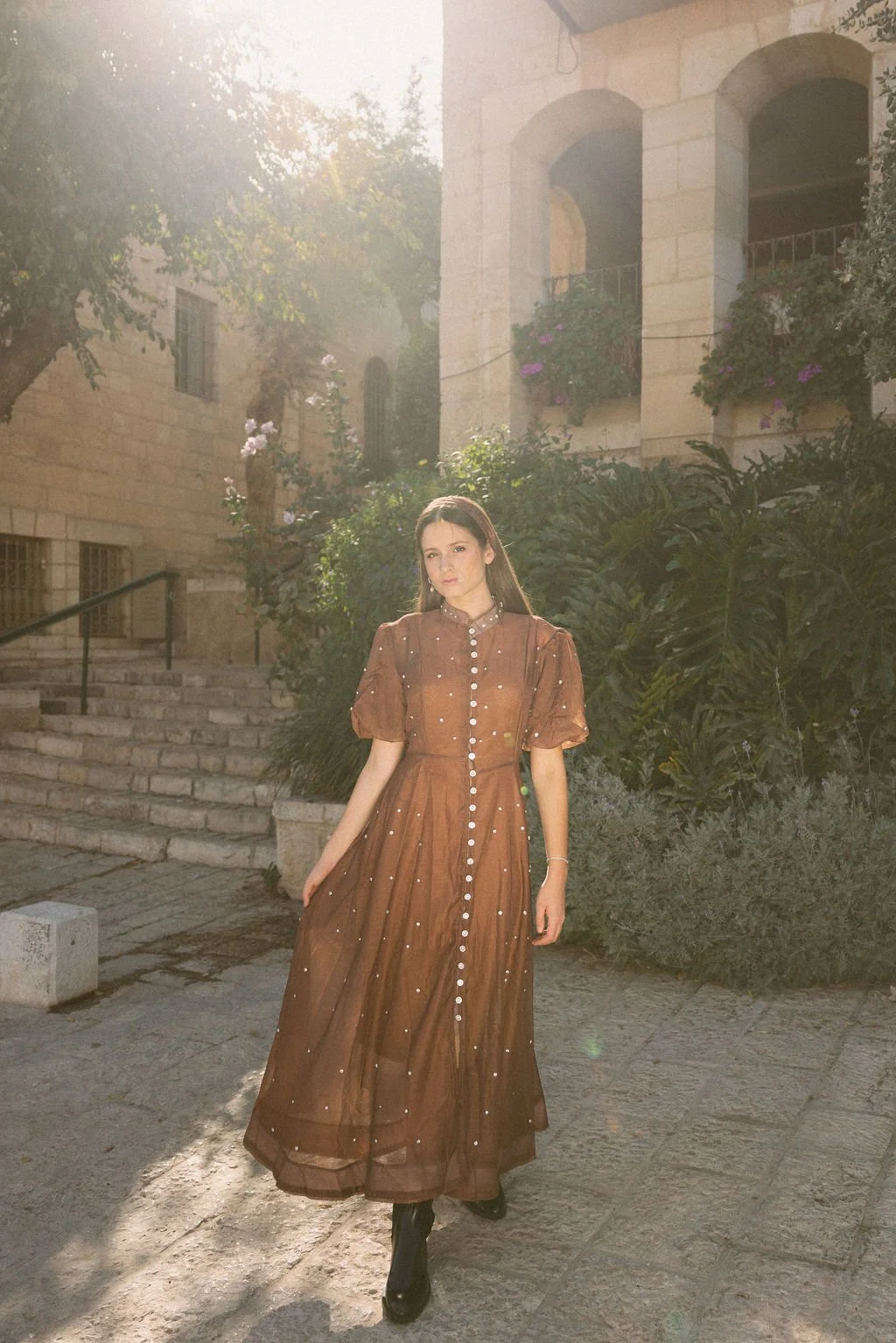 A woman in a brown, long dress with pearl details stands outdoors in a sunlit courtyard surrounded by greenery and stone buildings.
