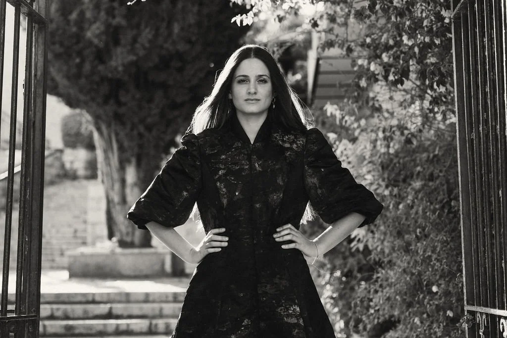 A woman with long hair standing outdoors with her hands on her hips, wearing a dark dress or coat, in front of a gated area with trees and steps in the background, in black and white.