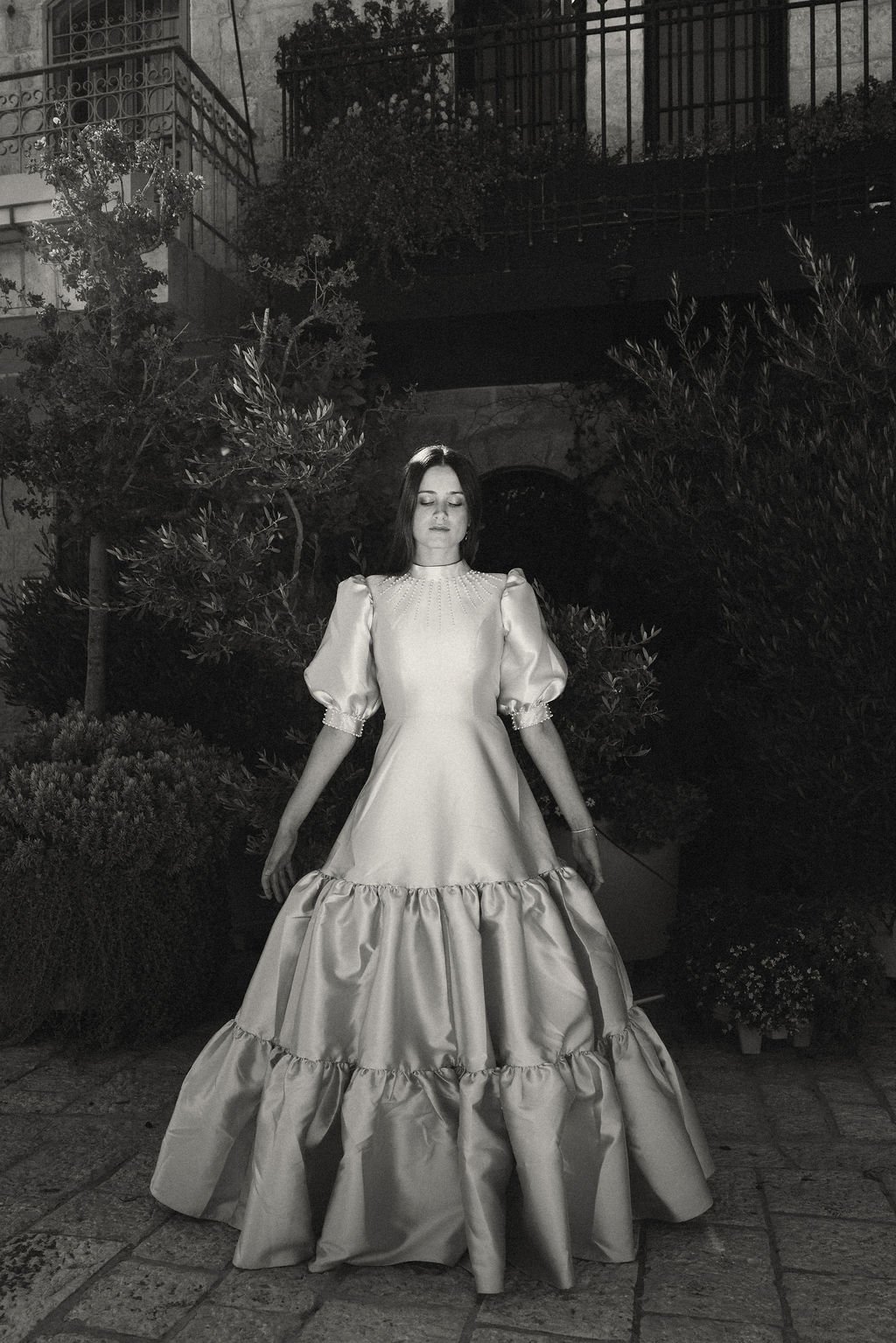 A woman in an elegant, vintage-style gown with puffed sleeves and a double-tiered skirt, standing outside at night with greenery and a building with balconies in the background.