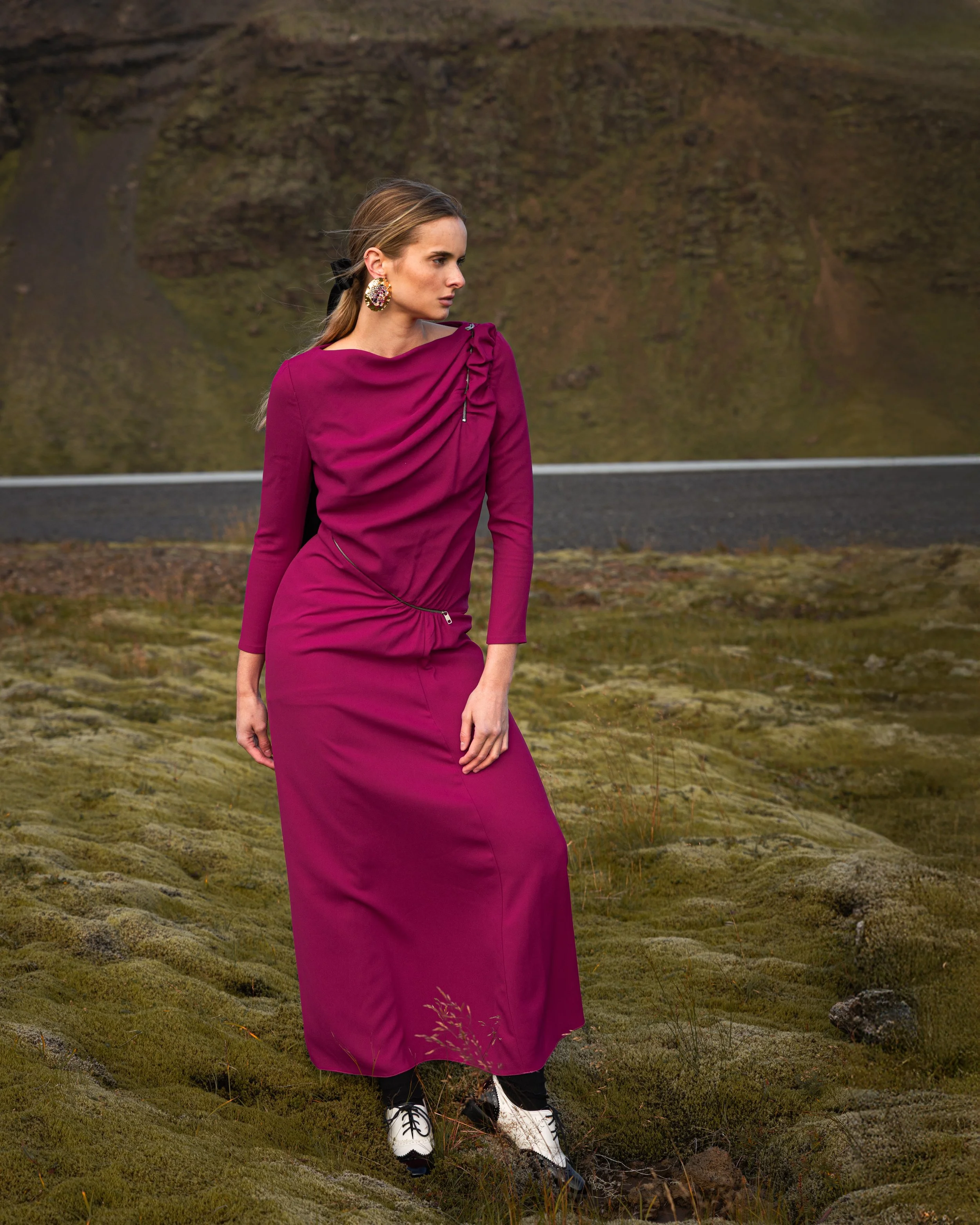 A woman in a long purple dress and white shoes with black laces stands outdoors on mossy terrain with mountains and a road in the background.