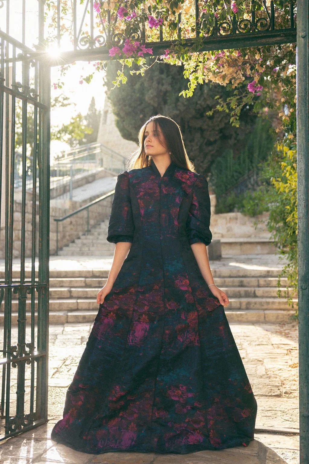 A woman in a long, dark floral dress stands under an iron gate with pink flowers, sunlight shining behind her, on a stone pathway with stairs.