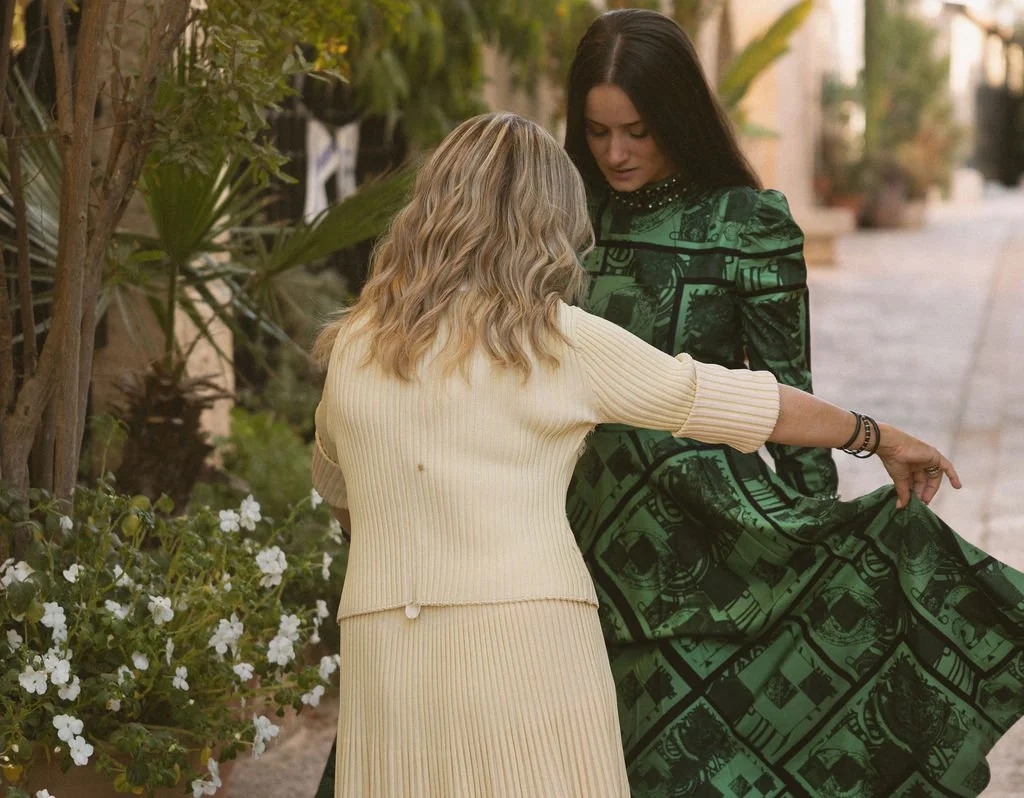 Two women are outdoors, one with blonde hair wearing a cream-colored ribbed dress pointing at a green patterned dress worn by the other woman with dark hair.
