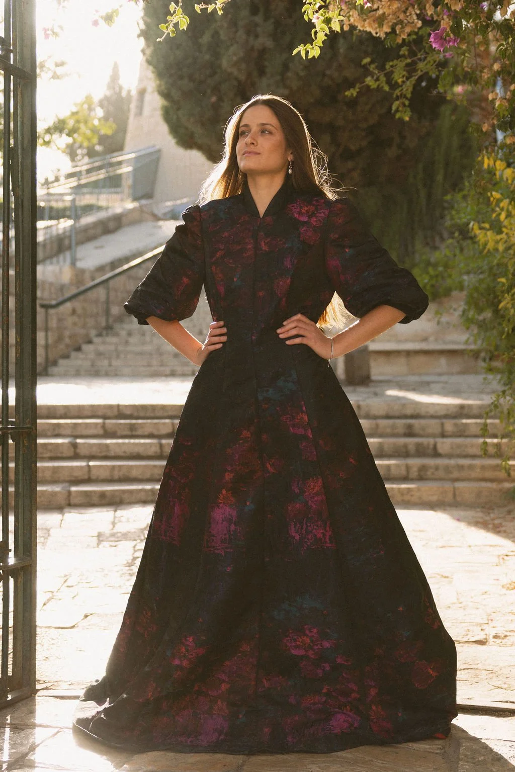 A woman in a long, dark floral dress with puffed sleeves stands outdoors with her hands on her hips, backlit by sunlight, near stone steps surrounded by greenery and pink flowers.