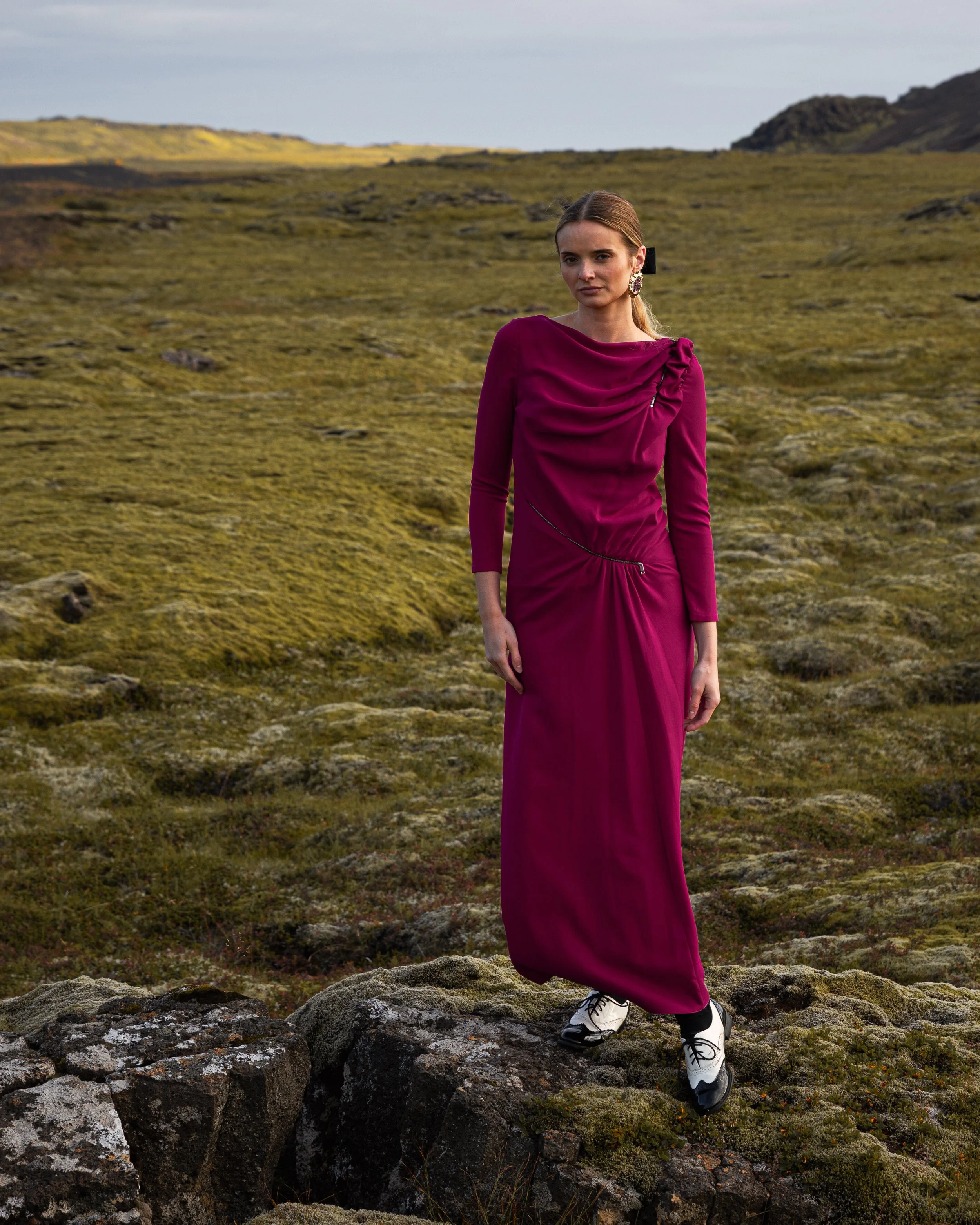 A woman in a magenta dress and white sneakers standing on rocks in an open grassy landscape with hills in the background.