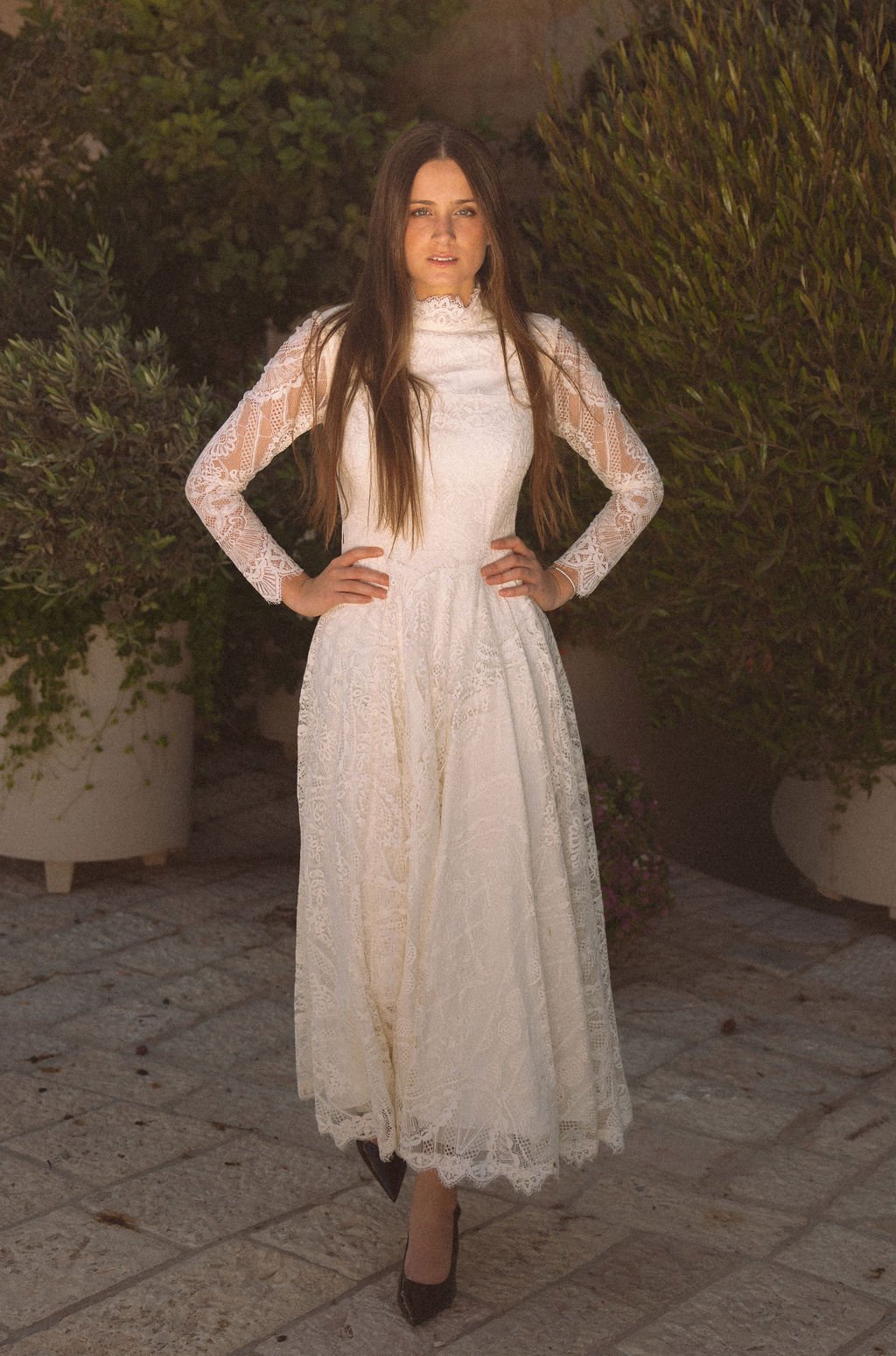 A woman wearing a long white lace dress with long sleeves, standing outdoors on a stone pathway surrounded by greenery.
