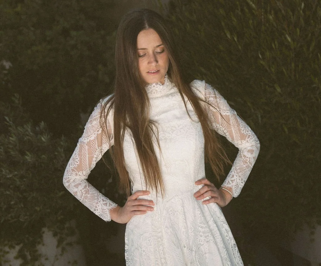 A young woman in a white lace dress with long sleeves and a high neckline, standing outdoors at night with her hands on her hips, looking down.