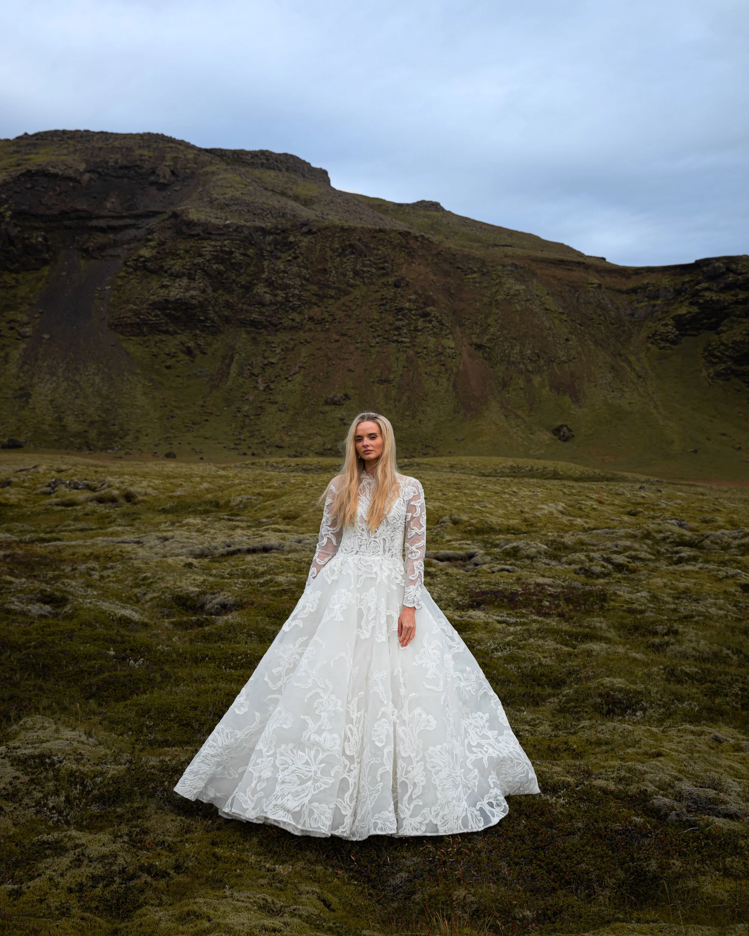 A woman in an elegant white lace wedding dress standing on mossy ground in a green, mountainous landscape.