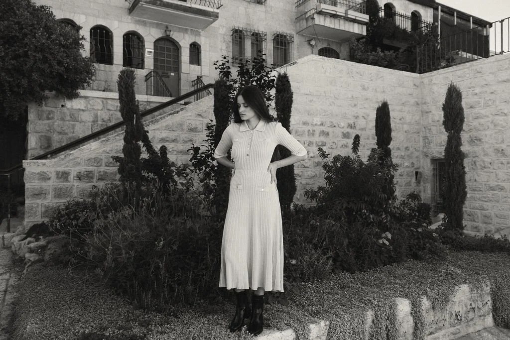 A woman in a long, pleated dress standing in a garden in front of a stone building with stairs and tall, slender trees.