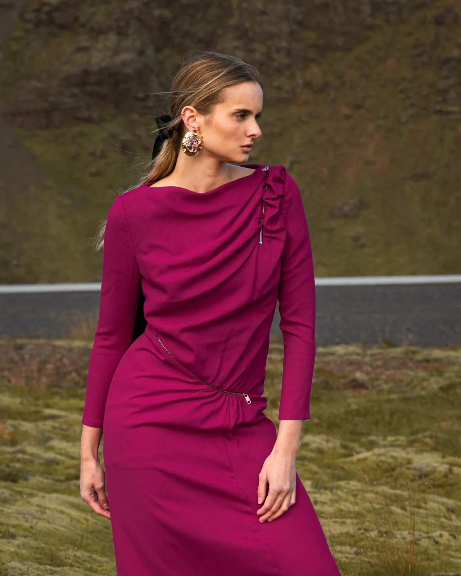A woman wearing a magenta long dress with draped fabric and zipper details, standing outdoors on a grassy, mountainous landscape.