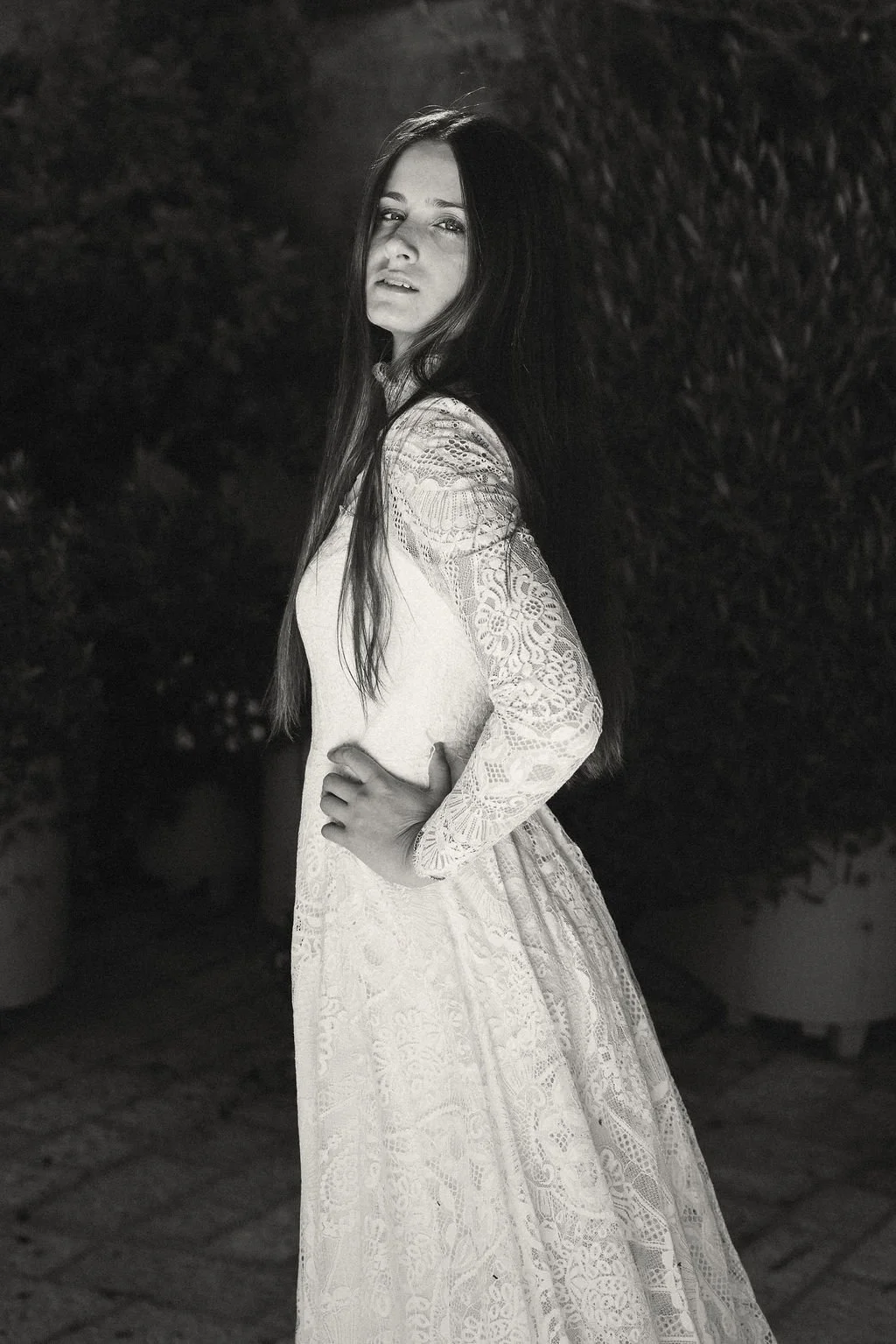 A woman with long hair wearing a lace long-sleeve dress, standing outdoors at night with her hand on her hip, looking at the camera.