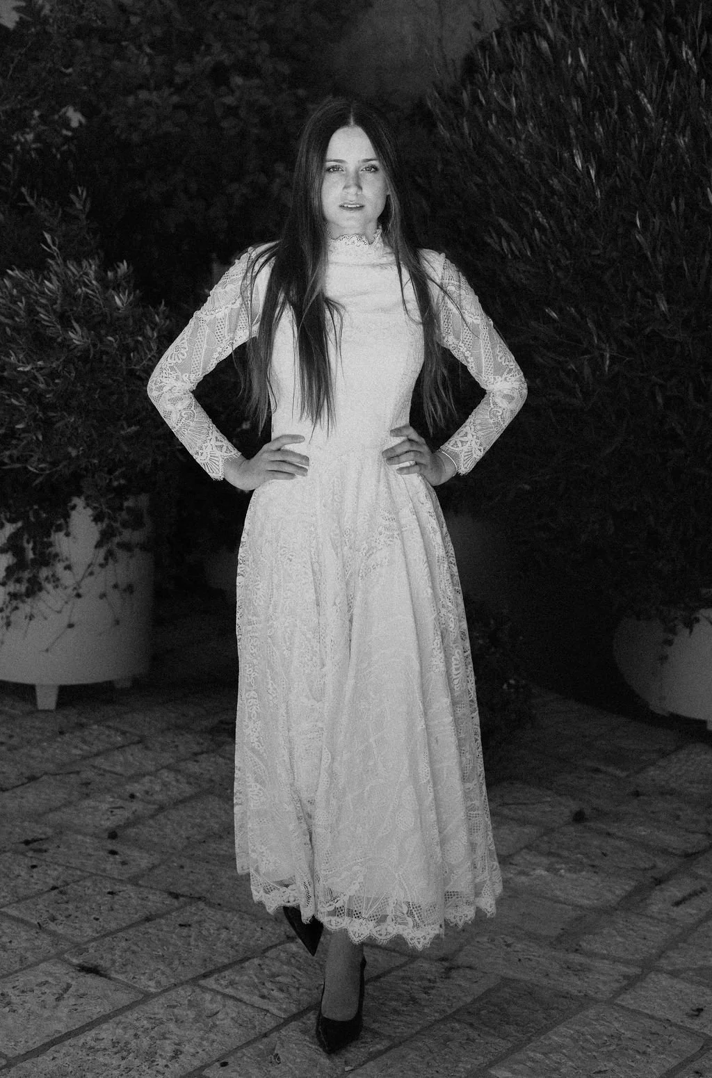 A woman in a long, lace dress standing outdoors with her hands on her hips, surrounded by plants.