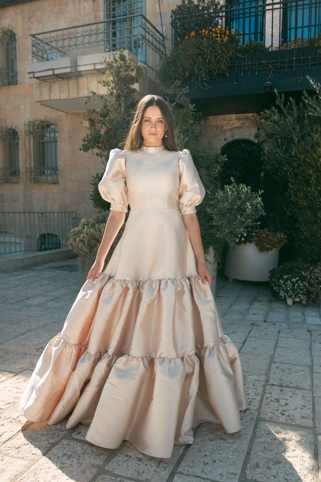 A woman wearing an elegant ivory satin evening gown with puffed sleeves and ruffled skirt standing outdoors on stone pavement with potted plants and a building with balconies in the background.