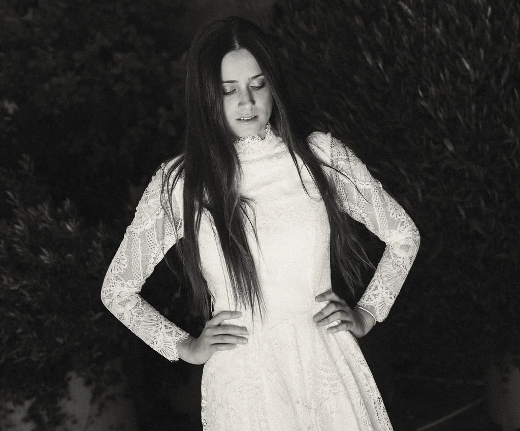 Black and white photo of a woman with long hair wearing a lace dress, standing outdoors at night with her hands on her hips, looking downward.