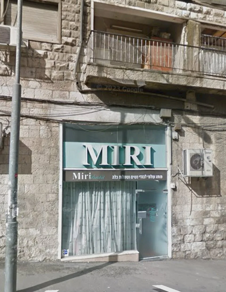Storefront with a sign that says 'MIRI' and bilingual text below, with curtains in the window and an air conditioning unit on the wall.