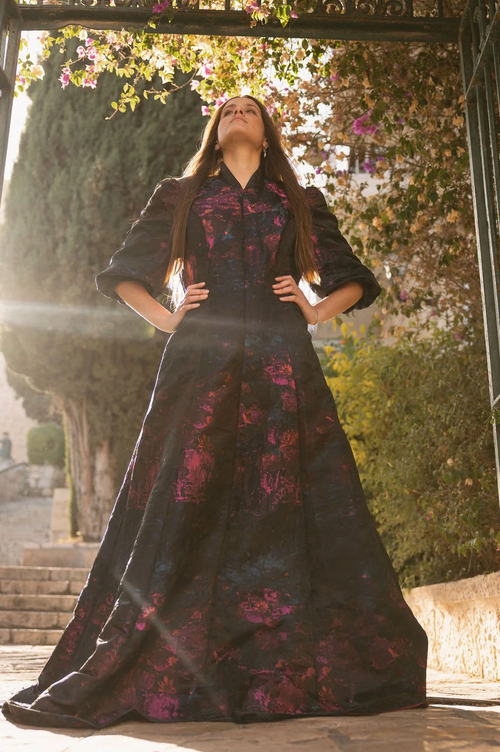 Woman in a long black dress with pink and purple floral patterns, standing outdoors under flowering bushes with her hands on her hips, looking upward.