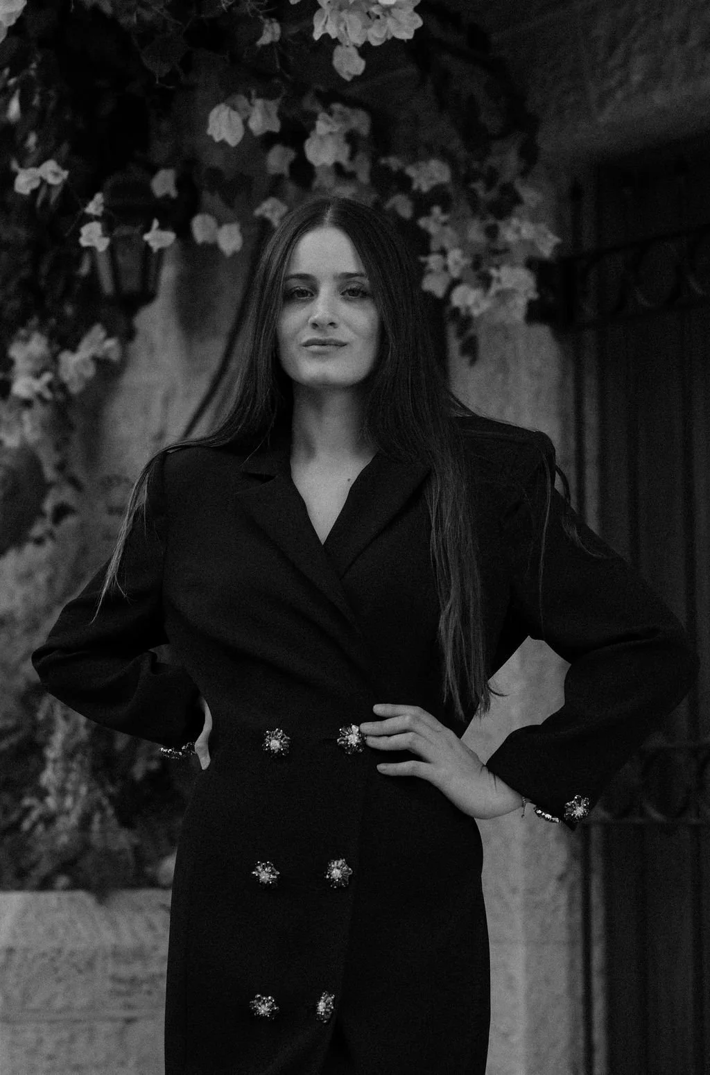 A woman with long hair poses with one hand on her hip, wearing a dark suit with decorative buttons, standing in front of a background adorned with flowers and foliage.