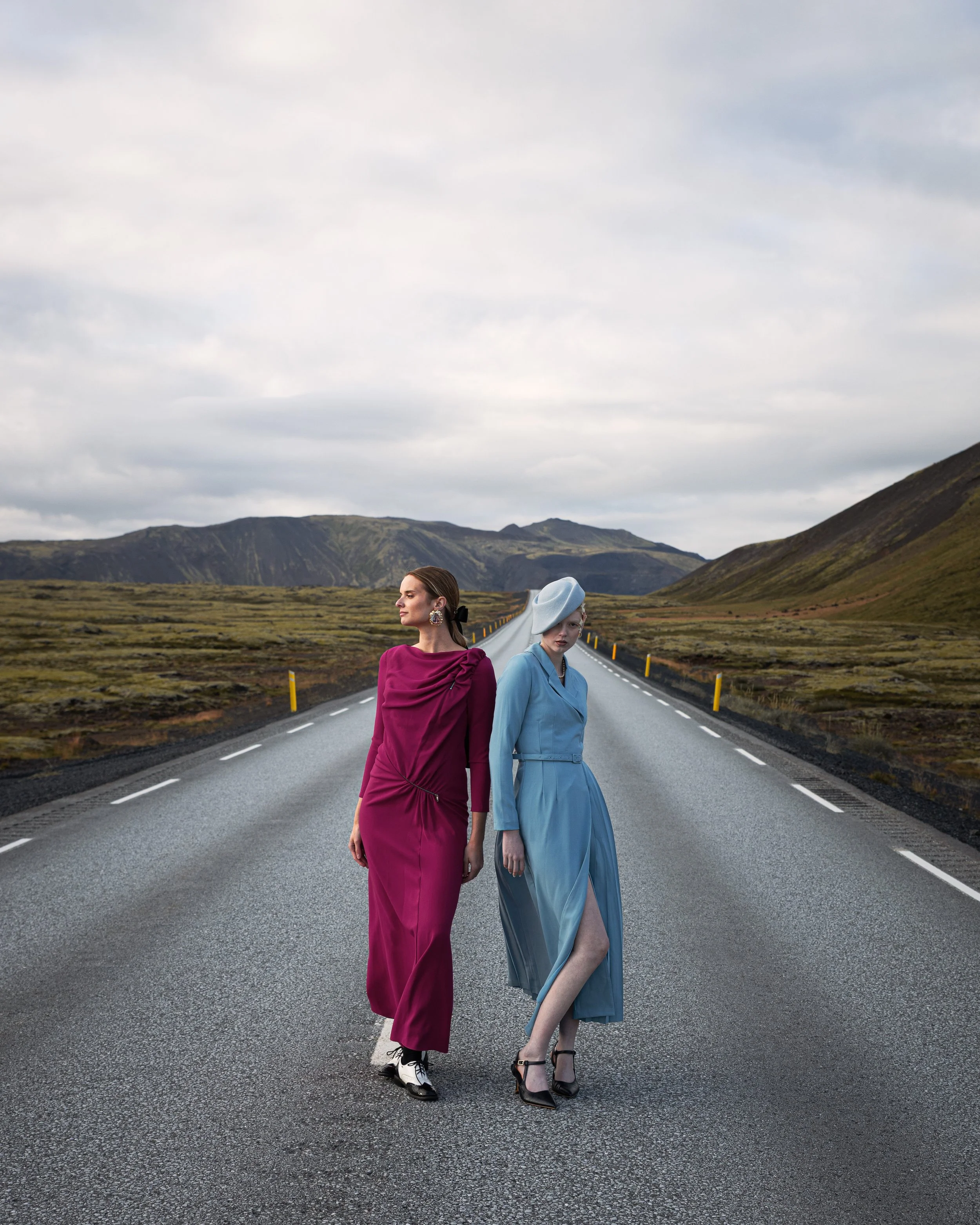 Two women in vintage dresses walking on an open road in a remote mountainous landscape.