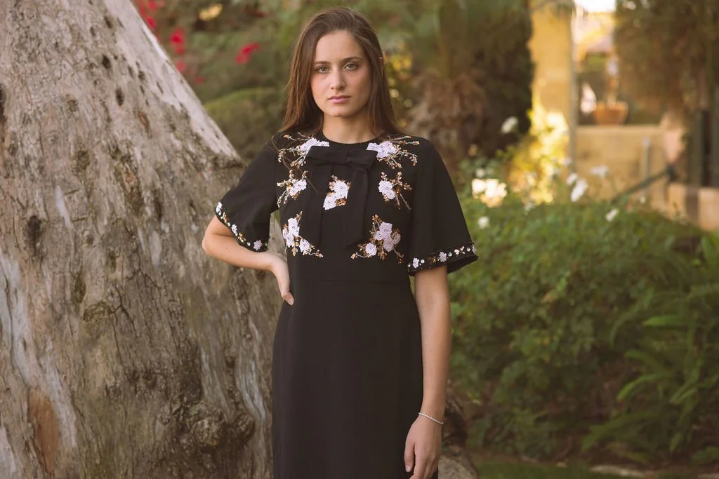 Young woman posing outdoors with trees and greenery in the background, wearing a black dress with floral embroidery and a bow detail on the chest.