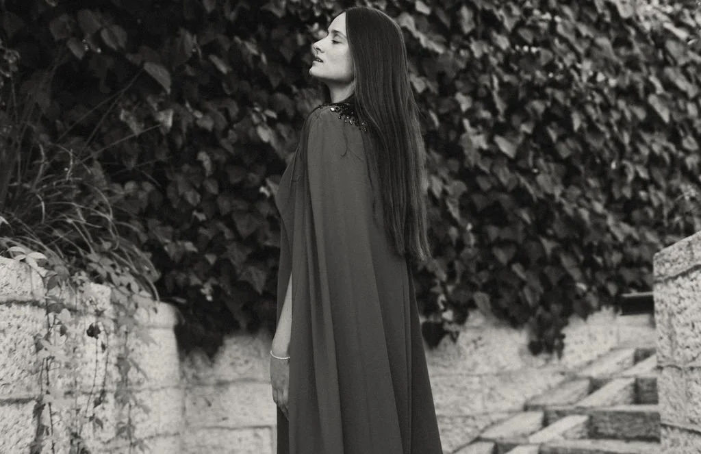 Woman with long dark hair in a graduation gown standing outdoors near stone steps and bushes.