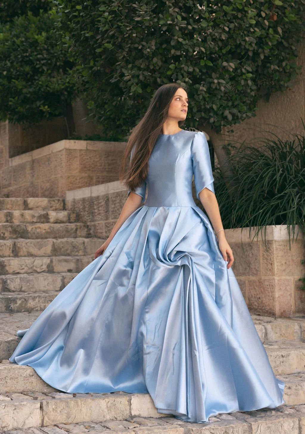 A woman in a shiny light blue ball gown stands on stone steps outdoors, with green bushes and a brick wall in the background.