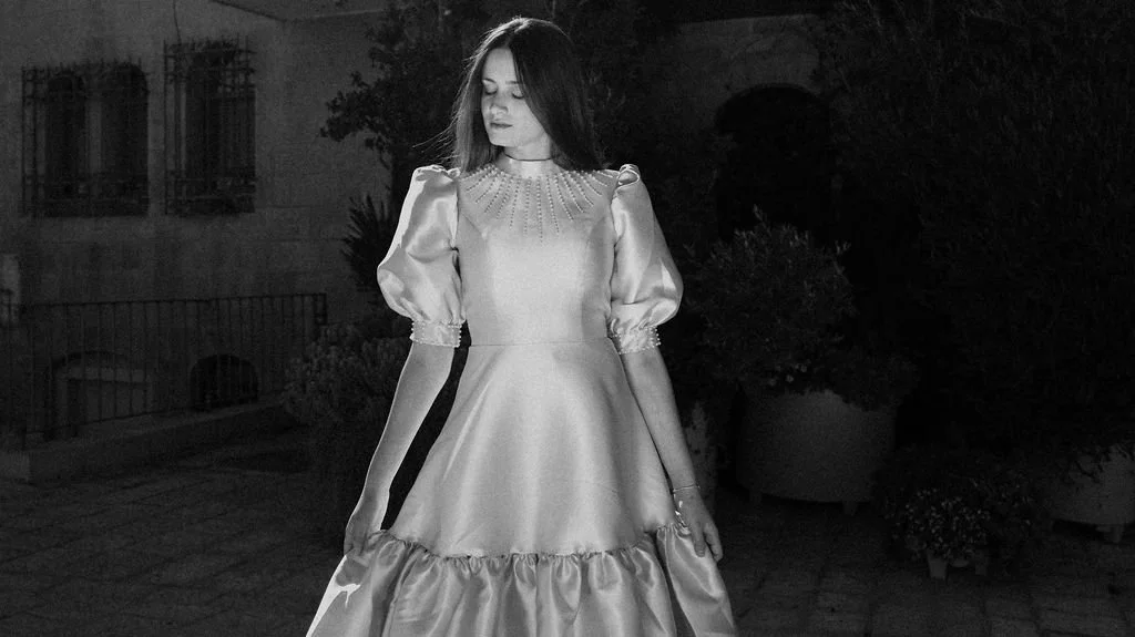 A woman in a vintage-style dress with puffed sleeves standing outdoors in a garden at night.