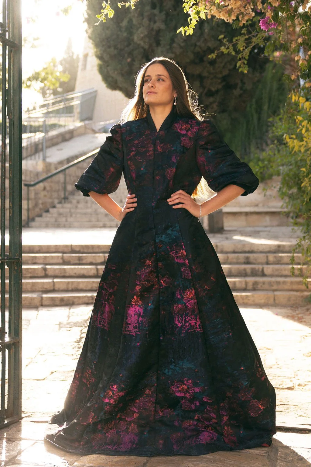 A woman in a long, dark floral dress standing outdoors on stone steps with hands on hips, backlit by sunlight and greenery.