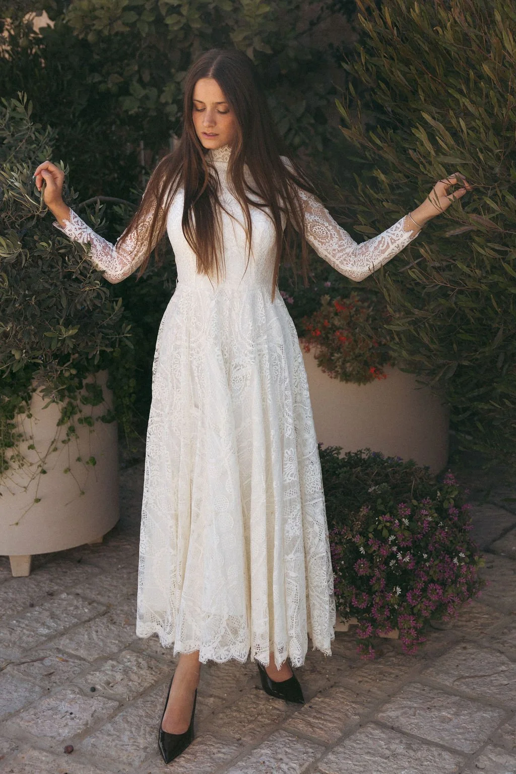 A woman in a long white lace dress standing outdoors among greenery and plants, with her eyes closed and arms slightly raised.