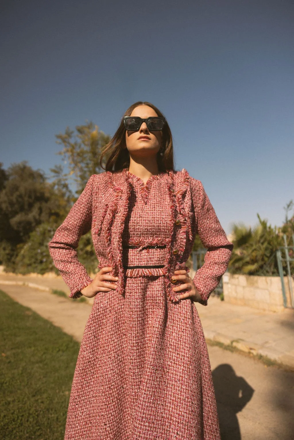 A woman standing outdoors wearing a pink tweed suit and oversized black sunglasses, with hands on her hips and a clear blue sky background.