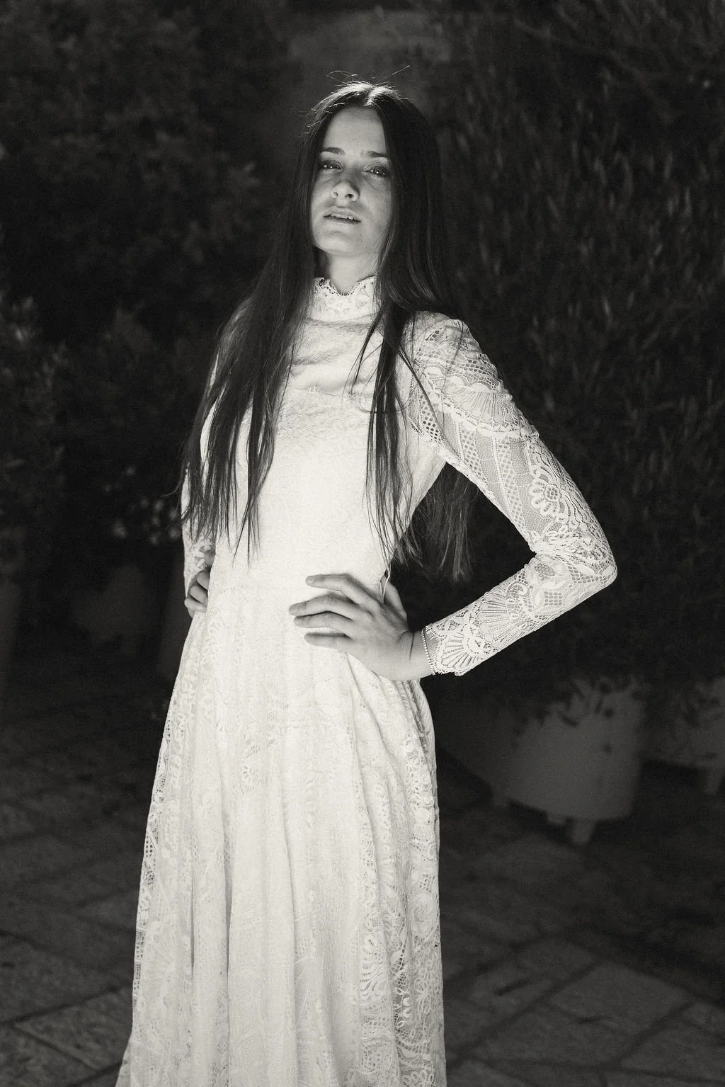 A black and white photo of a woman in a lace long-sleeve dress standing outdoors at night with trees in the background, one hand on her hip and the other on her waist.
