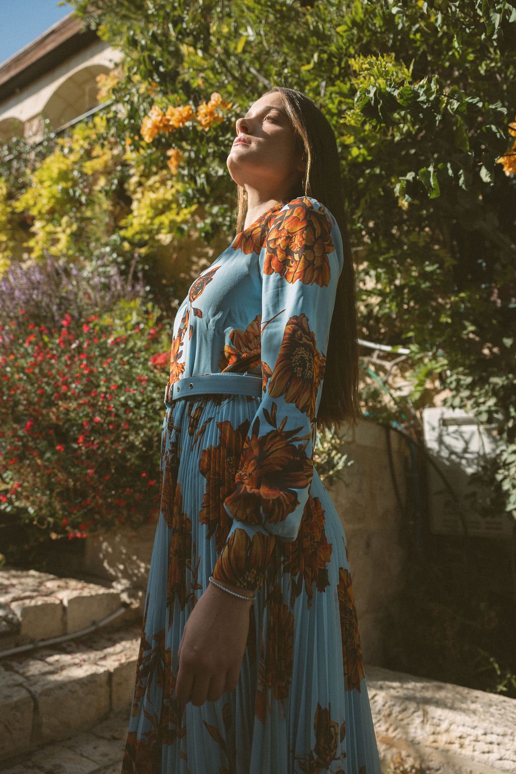 A woman with long dark hair standing outdoors in sunlight, wearing a blue dress with orange floral patterns, in a garden with green foliage and flowering bushes.