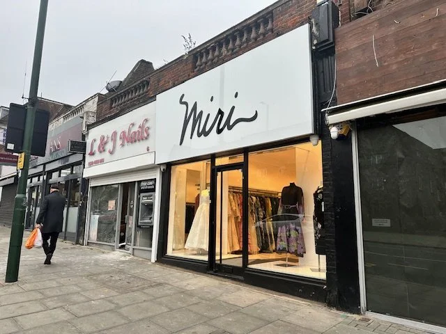 Street view of a retail store named 'Mimi' selling wedding dresses, with a man walking by and an adjacent business named 'L & L Nails'.