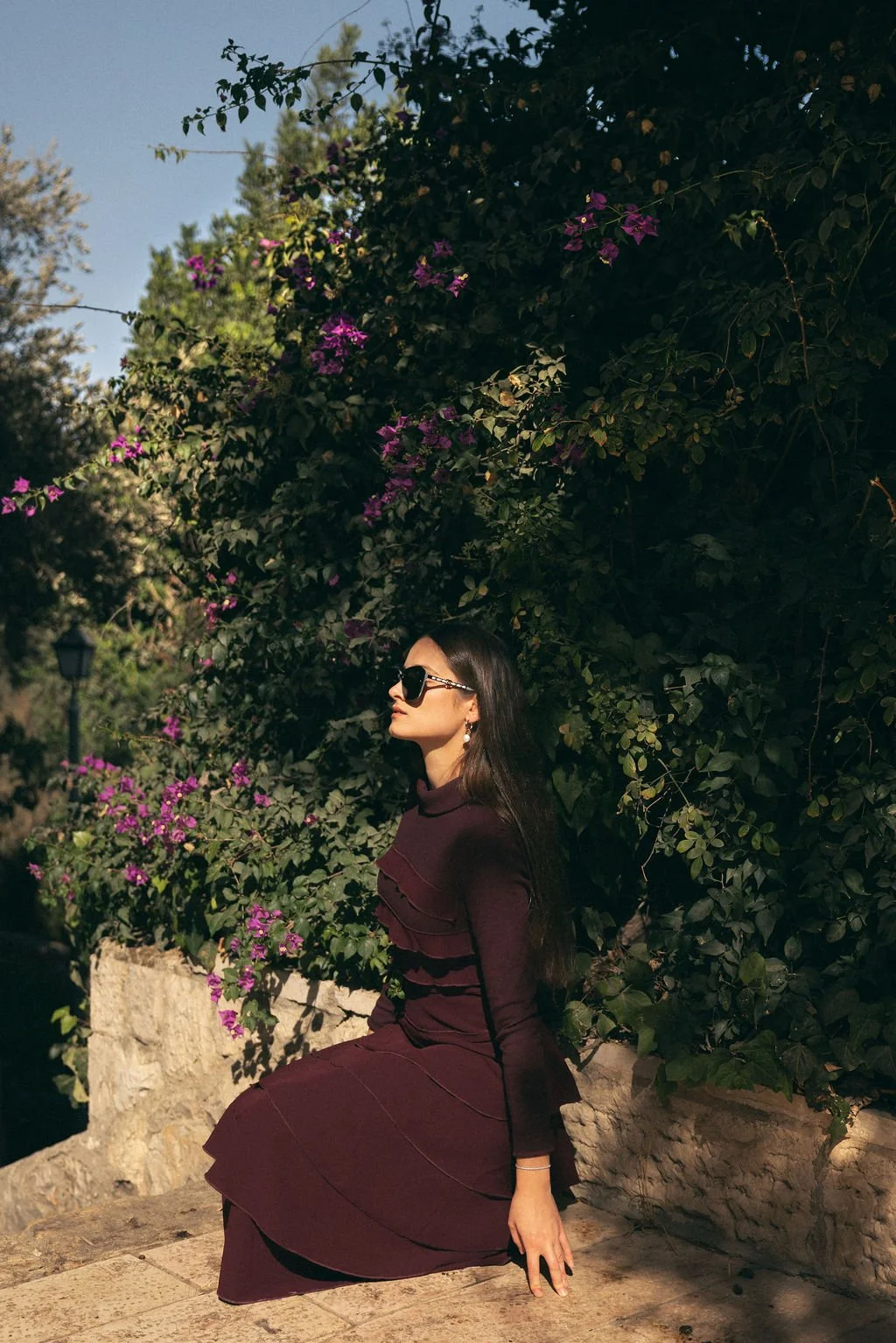 A woman with long dark hair wearing dark sunglasses and a burgundy dress with ruffles, sitting on a stone ledge next to green shrubbery and purple flowers, in sunlight.