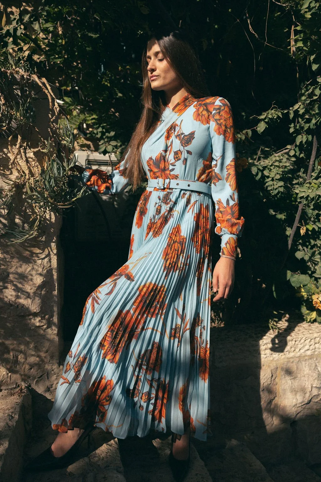 A woman wearing a light blue, long-sleeved floral dress with orange and brown flowers, standing outdoors with greenery and rocks behind her.