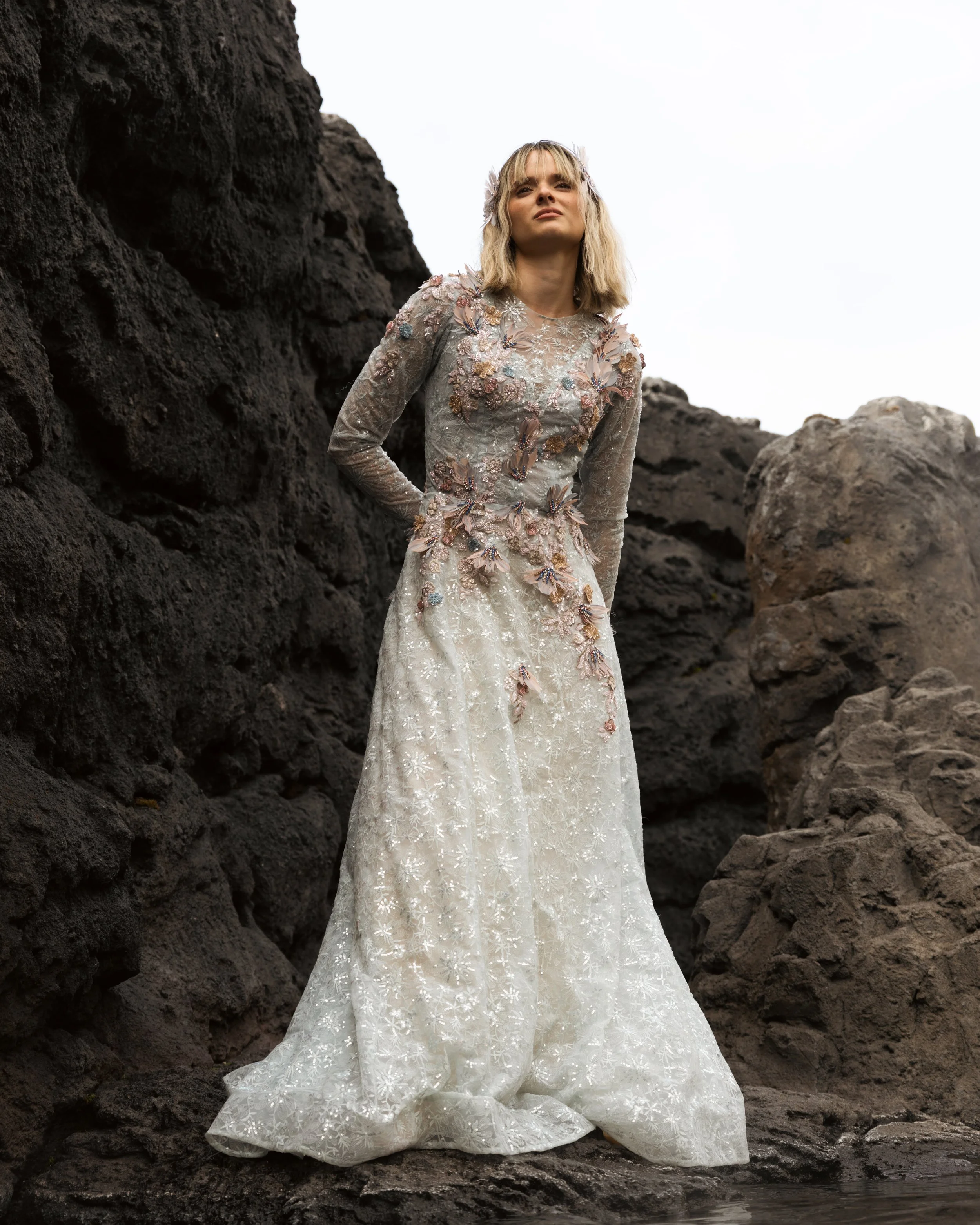 A woman wearing a long, ornate wedding dress standing among dark volcanic rocks.
