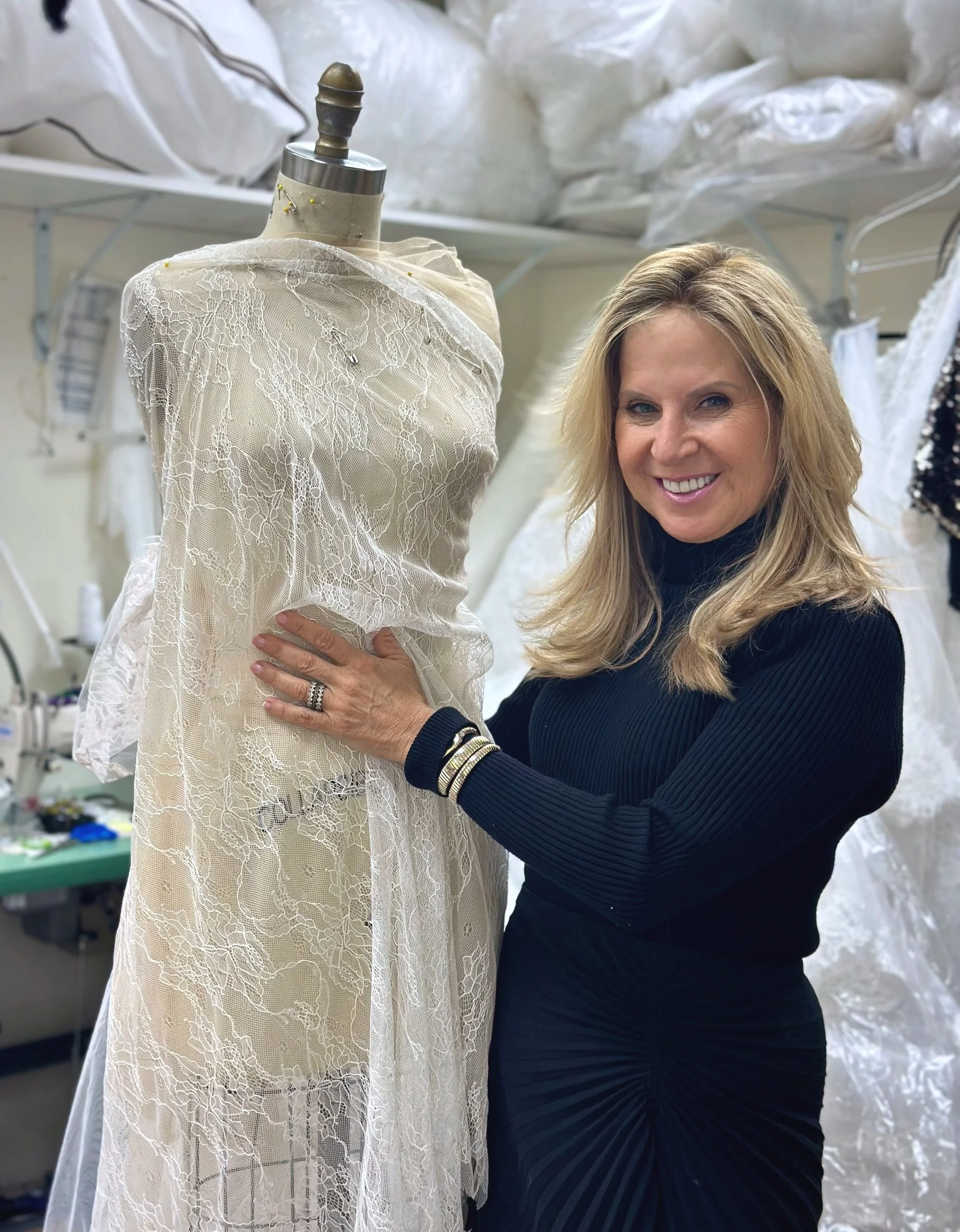 A woman with blonde hair smiling and wearing a black long-sleeve top stands next to a dress form draped with white lace fabric. The background shows a workspace with fabrics and sewing materials.