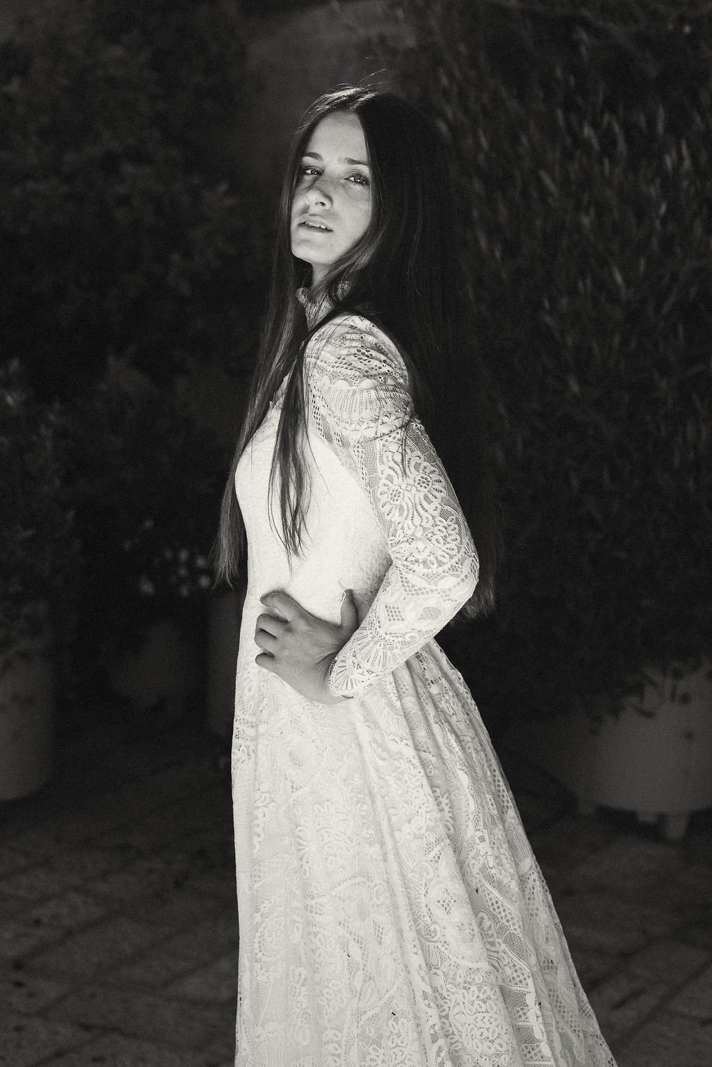 A woman with long dark hair wearing a lace long-sleeve dress, standing outdoors at night with a dark, leafy background.