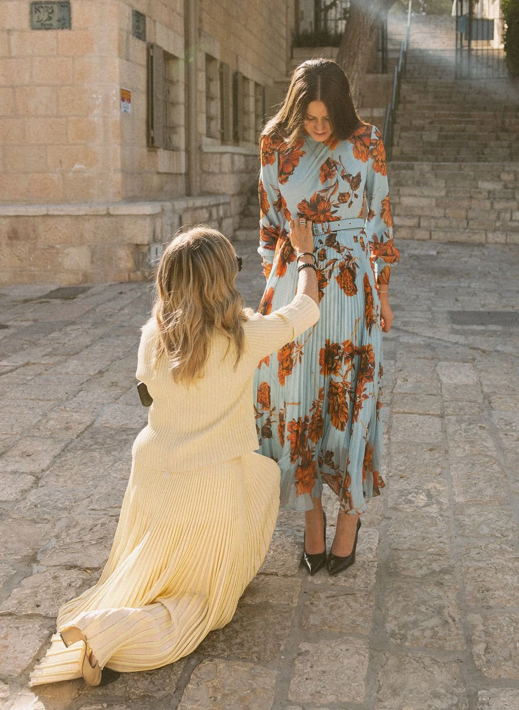 Woman in floral dress standing while another woman kneels, touching her dress, on a cobblestone street outside.