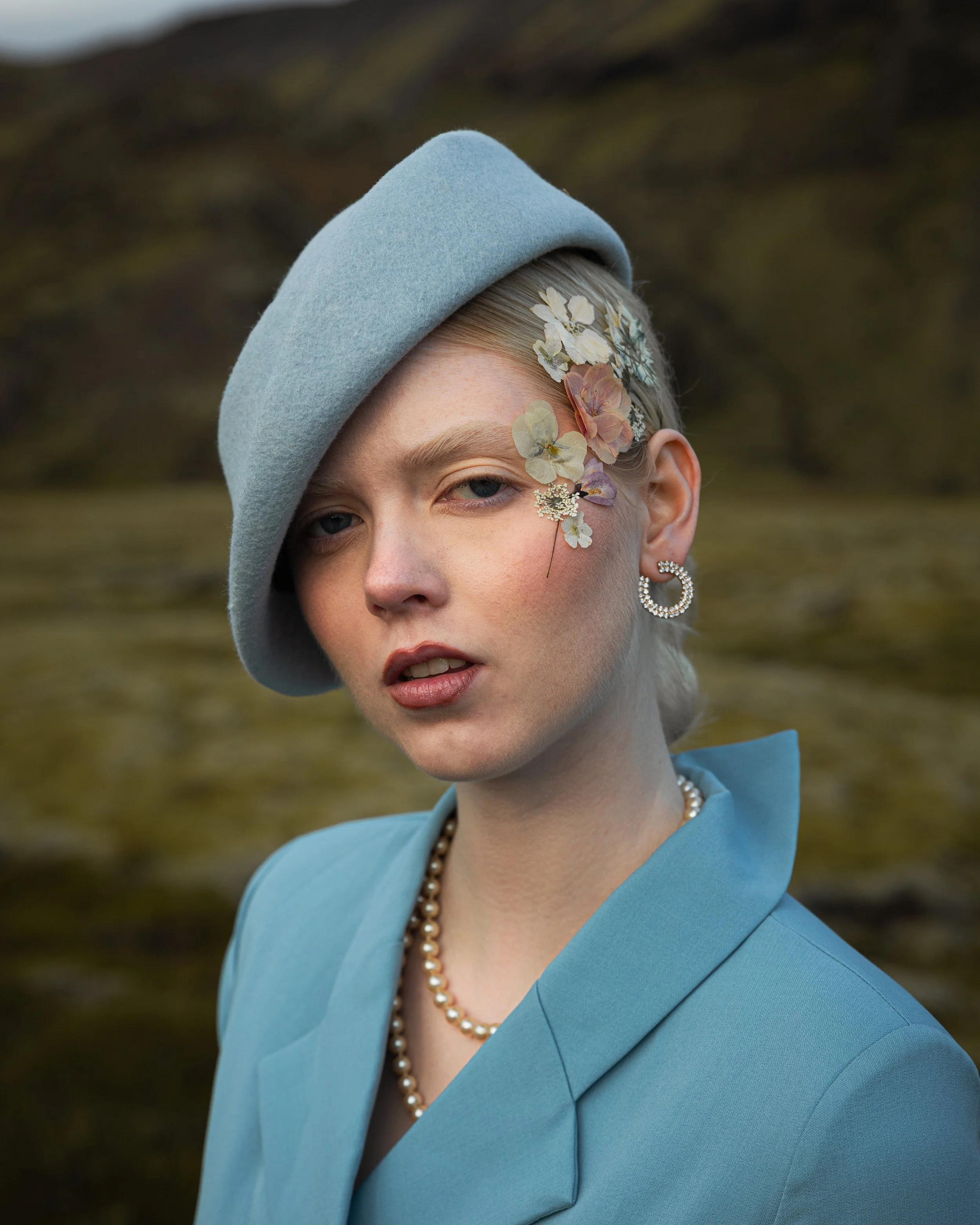 A young woman with blonde hair styled back, wearing a light blue coat, a gray beret, pearl necklace, and pearl earrings. She has decorative flowers and leaves on her face near her right eye, with a neutral expression against a natural outdoor backgro