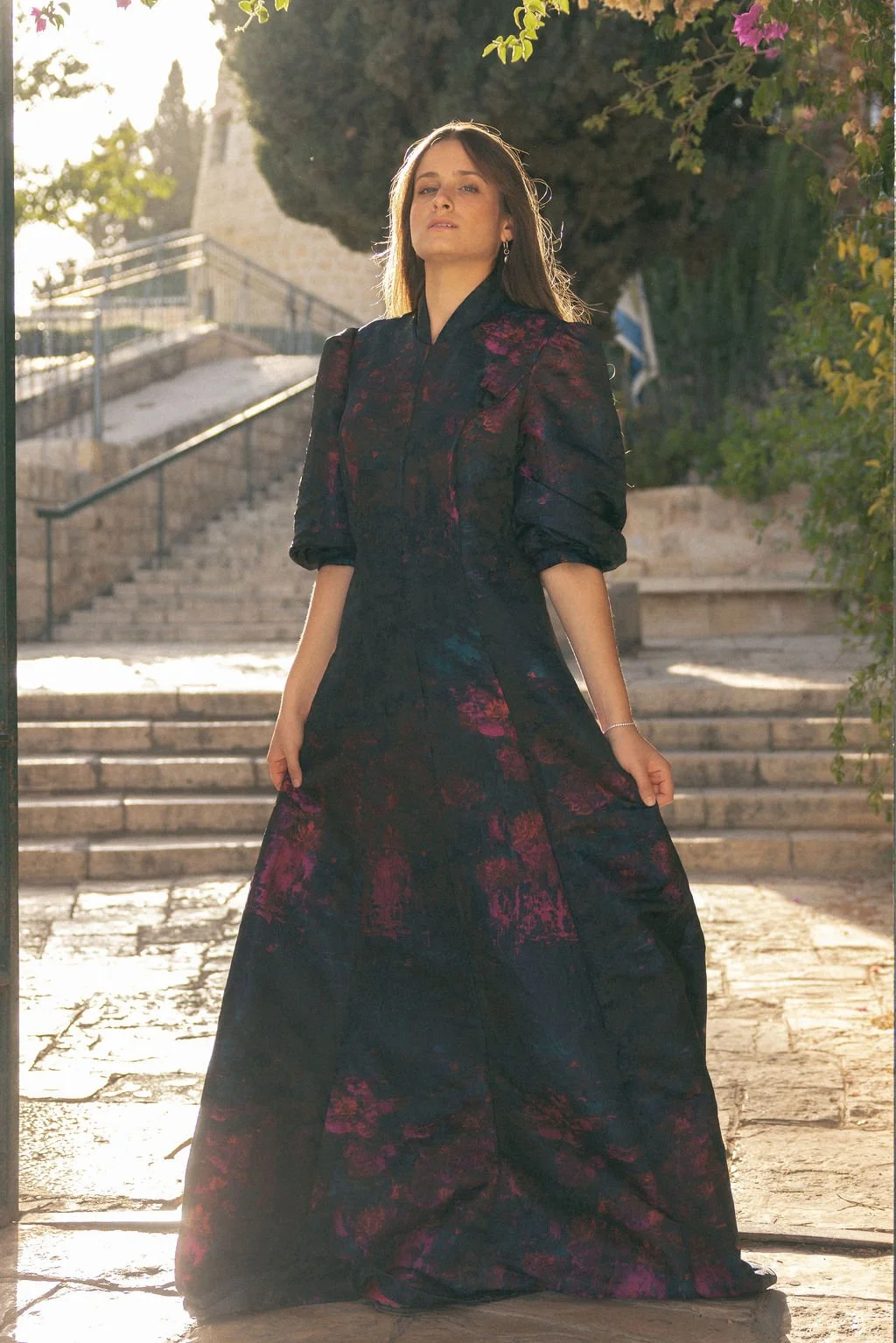 A woman standing outdoors on stone steps, wearing a long dark floral dress with puffed sleeves, backlit by sunlight, with greenery and flowering plants around her.