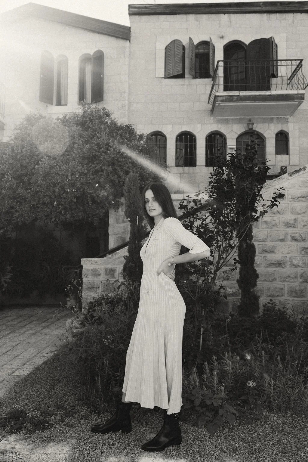 A woman in a long, light-colored striped dress and black boots standing outdoors near a garden with bushes and trees. In the background, there is a stone building with open window shutters, a balcony, and a staircase.